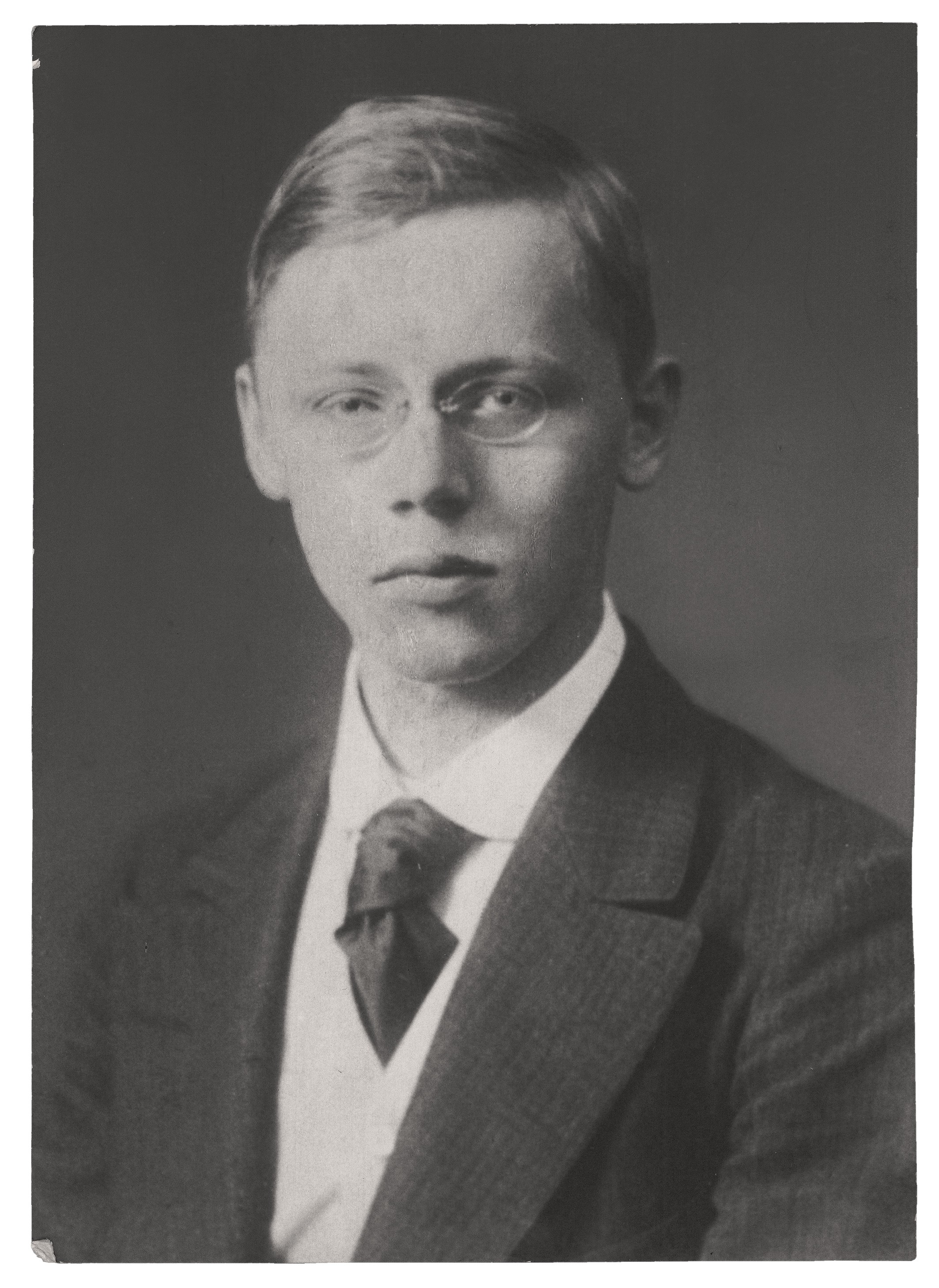Das Schwarz-Weiß-Porträt zeigt die Brustansicht von Hans Kollwitz als Student, mit schmalem Gesicht und ernsthaftem, melancholischem Blick. Er trägt eine Brille mit runden Gläsern und sein kurzes, helles Haar ist glatt zur Seite gescheitert. Seine Kleidung besteht aus einem dunklen, fein gestreiften Sakko über einer hellen Weste und einem weißen Hemd mit einer kunstvoll verknoteten, dunklen Krawatte.