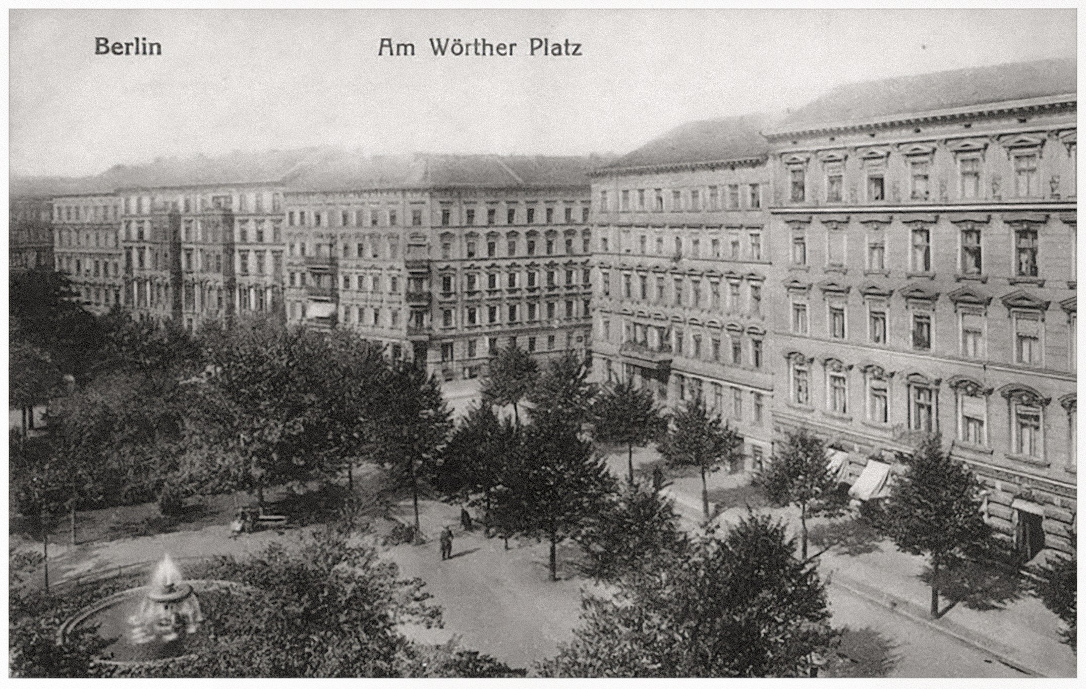 Das Bild zeigt eine historische Schwarz-Weiß-Postkarte vom Wörther Platz in Berlin, auf der ein belebter Stadtplatz mit einem zentralen Springbrunnen und dichtem Baumbestand zu sehen ist. Im Hintergrund ragen prachtvolle, mehrstöckige Wohnhäuser mit reich verzierten Fassaden empor, die typisch für die Architektur der Berliner Gründerzeit sind. Die Szene wirkt friedlich und fängt die Atmosphäre des städtischen Lebens in einer vergangenen Ära ein.