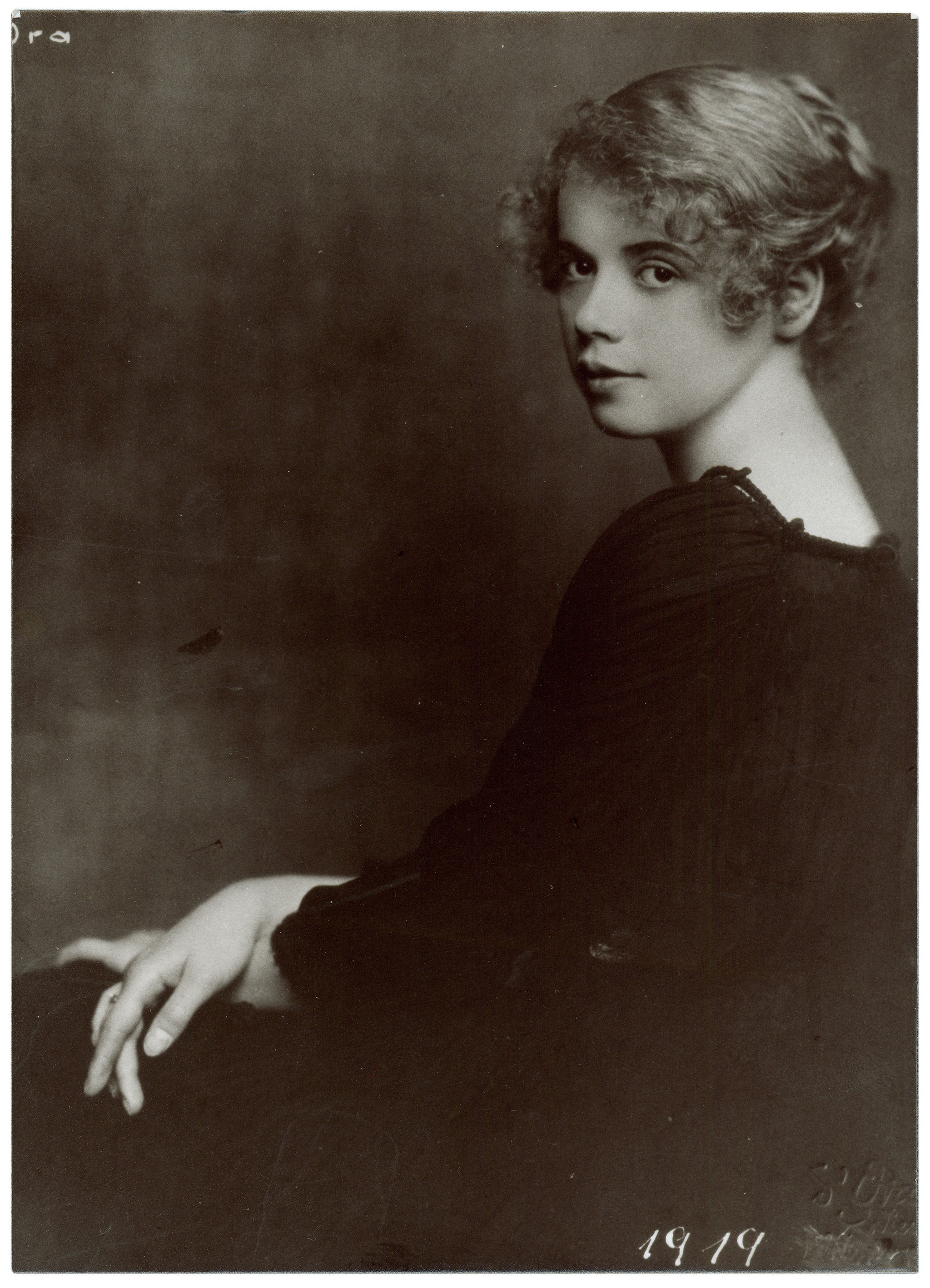 Dieses Porträt von 1919 zeigt Katharina Stern (1897–1984), alias Katta Sterna, als junge Frau in einer klassischen Seitenansicht, wobei sie ihren Kopf sanft über die Schulter zum Betrachter dreht. Ihr helles, lockiges Haar ist elegant hochgesteckt, was ihre feinen Gesichtszüge und ihren ruhigen, klaren Blick betont. Sie trägt ein dunkles Oberteil mit einer feinen Raffung am Ausschnitt, das sich deutlich von dem weichen, schattierten Hintergrund abhebt.