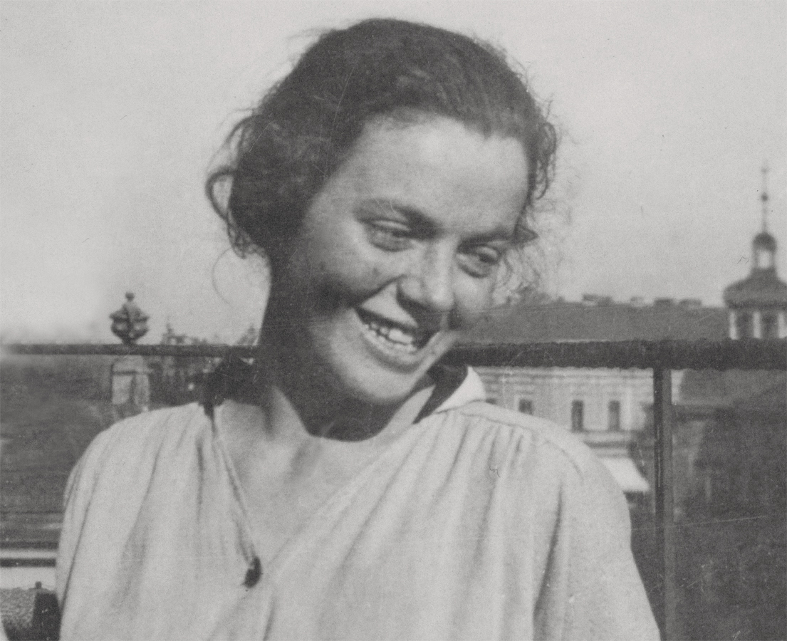 Das Bild zeigt eine Schwarz-Weiß-Fotografie von Regula Stern (1894–1978) als junge Frau, die in einem hellen, locker fallenden Oberteil vor einem Geländer im Freien steht. Sie blickt mit leicht geneigtem Kopf lächelnd nach unten, während ihr dunkles, leicht lockiges Haar vom Wind ein wenig zerzaust wird. Im unscharfen Hintergrund sind die Umrisse städtischer Gebäude und ein Turm unter hellem Himmel zu erkennen.