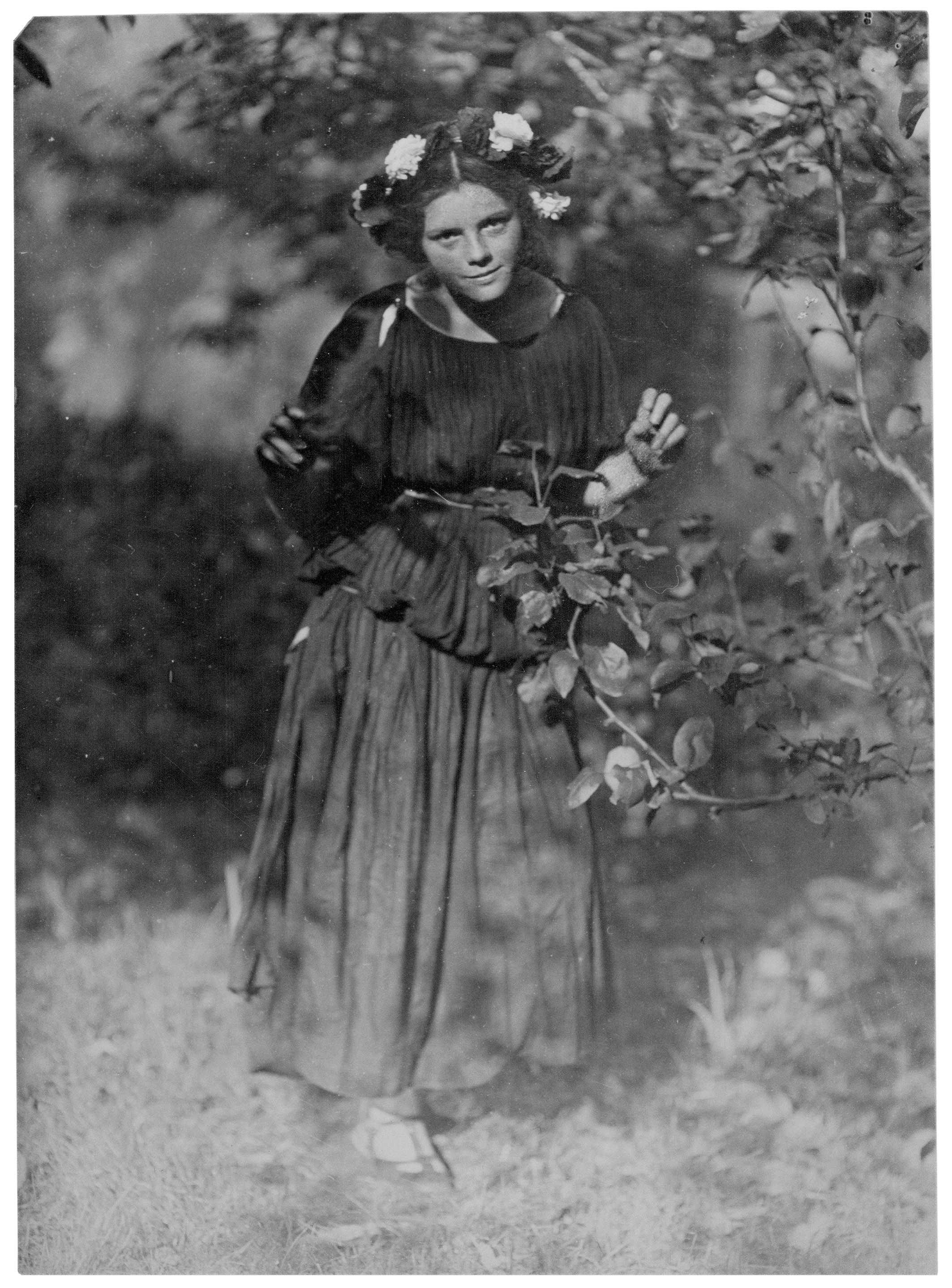 Das historische Schwarz-Weiß-Foto von 1912 zeigt Hanna Stern (1896–1988), alias Johanna Hofer, als junges Mädchen in einer natürlichen Umgebung, vermutlich einem Garten. Es trägt ein dunkles, locker fallendes Gewand mit einem weiten Rock und hat einen auffälligen Blumenkranz mit hellen und dunklen Blüten im Haar. In der Bildmitte steht das Mädchen aufmerksam blickend, umgeben von Laub und Zweigen, die im Vordergrund teilweise unscharf ins Bild ragen.