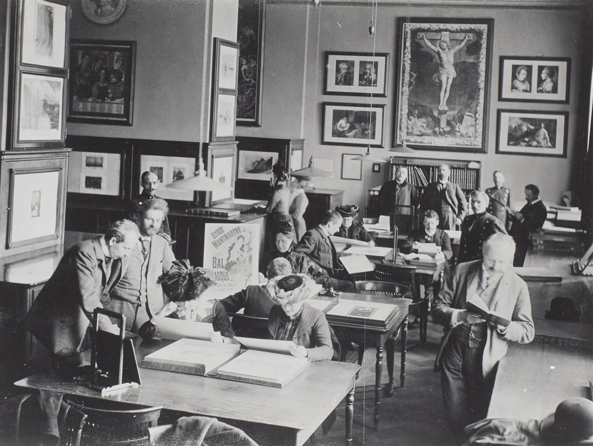 Die Fotografie von 1913 zeigt den Studiensaal des Kupferstich-Kabinetts im Museumsbau Gottfried Sempers, einen belebten, historischen Lesesaal oder eine Studiensammlung, in dem zahlreiche Personen in formeller Kleidung an großen Holztischen konzentriert über Mappen und Büchern arbeiten. Die Wände sind dicht an dicht mit gerahmten Grafiken, Zeichnungen und einem großen Kruzifix-Gemälde behängt, was die Atmosphäre eines wissenschaftlichen Instituts oder einer Kunstsammlung unterstreicht. Im Vordergrund stehen und sitzen Männer und Frauen in zeittypischer Garderobe, wie Sakkos und ausladenden Hüten, während das einfallende Licht eine kontrastreiche, ruhige Arbeitsstimmung erzeugt. Im Hintergrund rechts ist Max Lehrs zu sehen. 