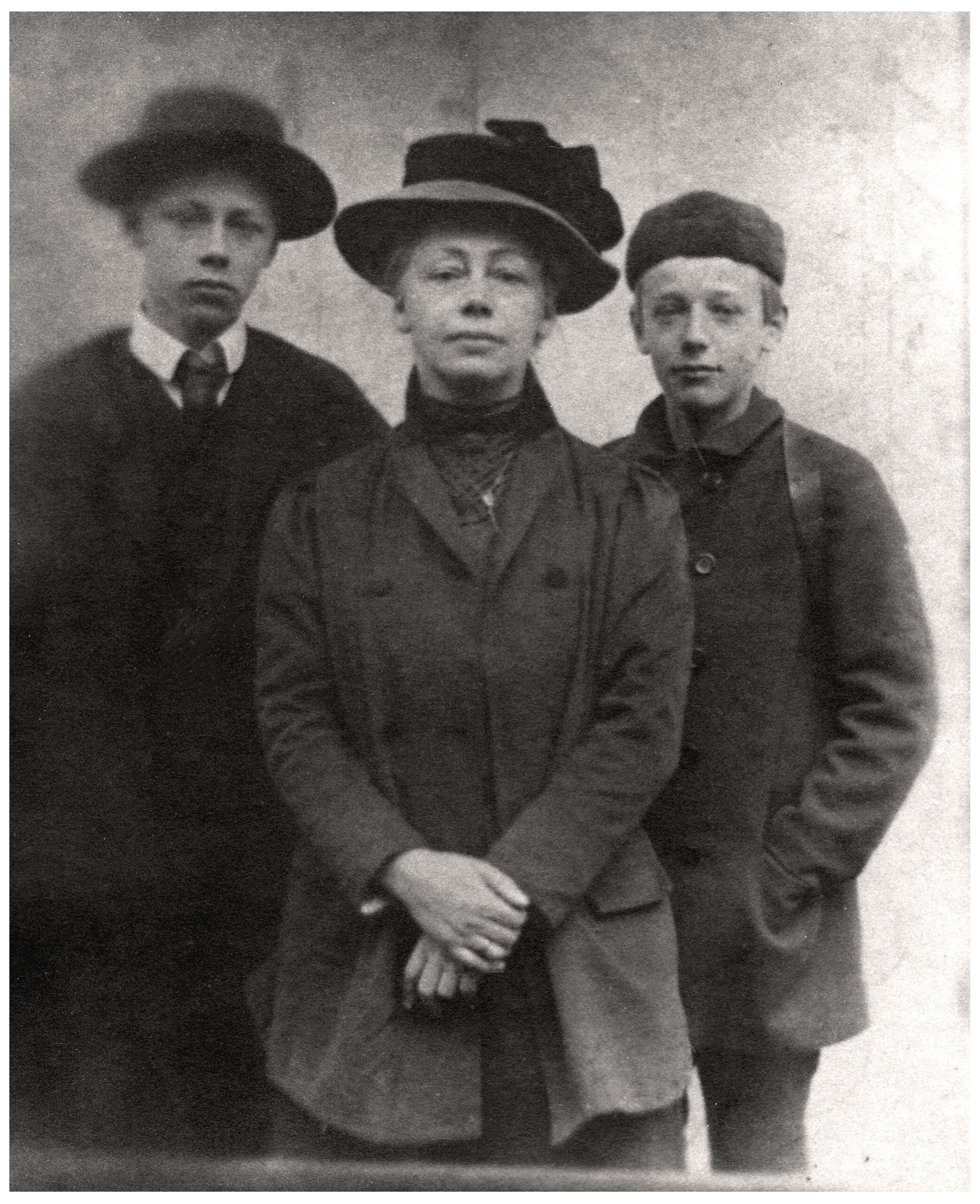 Dieses Schwarz-Weiß-Foto zeigt die Künstlerin Käthe Kollwitz in der Bildmitte, flankiert von ihren beiden Söhnen Hans und Peter. Alle drei Personen blicken ernst direkt in die Kamera und tragen dunkle, zeitgenössische Kleidung sowie markante Hüte. Die Komposition wirkt förmlich und ruhig, wobei der Fokus auf der Verbundenheit der kleinen Gruppe vor einem schlichten, hellen Hintergrund liegt.