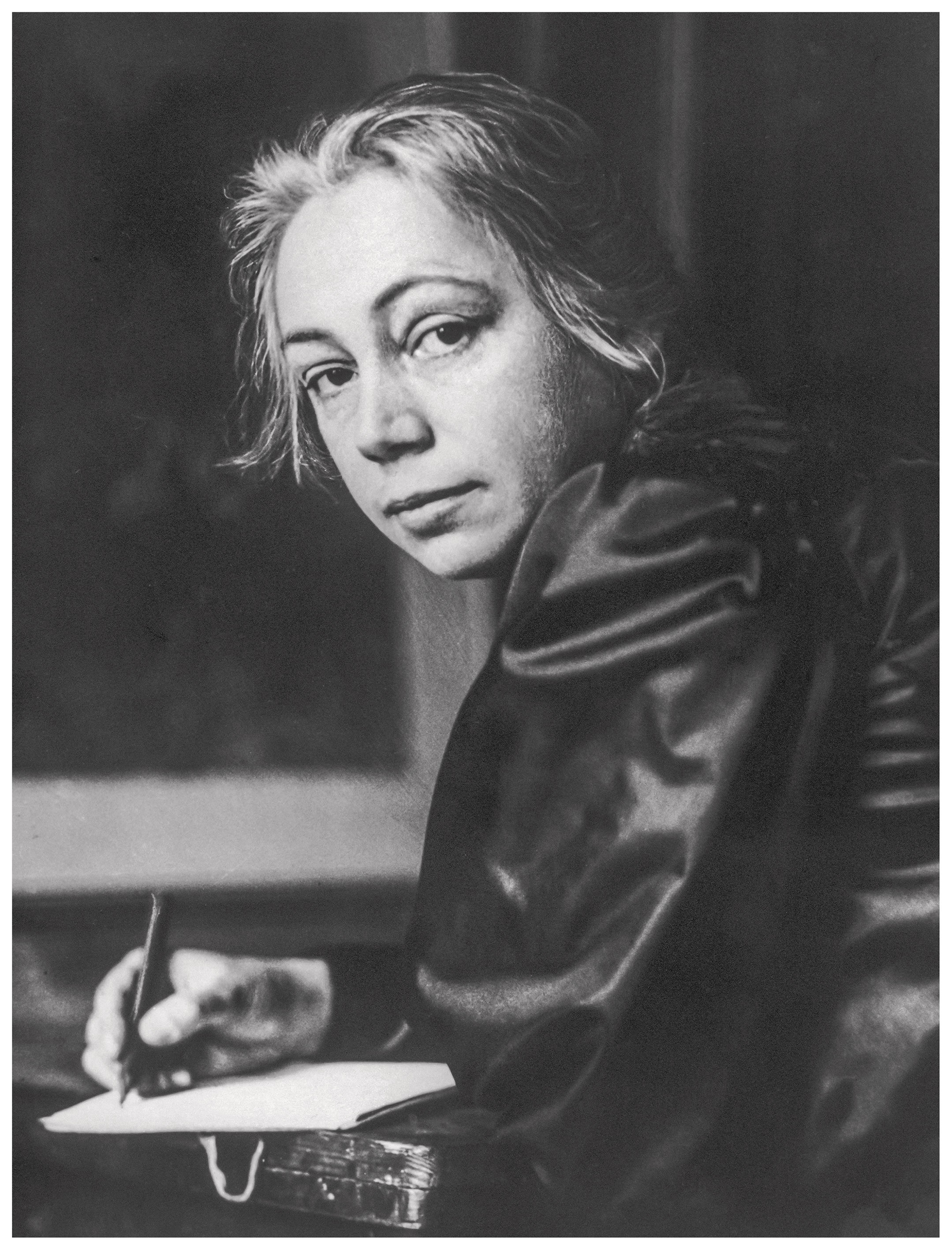 Das Bild zeigt eine Schwarz-Weiß-Fotografie der Künstlerin Käthe Kollwitz, die an einem Tisch sitzt und den Betrachter mit einem ernsthaften, nachdenklichen Blick direkt ansieht. Sie hält einen Stift in der Hand und scheint gerade beim Schreiben oder Zeichnen auf einem Papierblock unterbrochen worden zu sein. Ihr dunkles Oberteil glänzt seidig im Licht, während ihr Haar schlicht nach hinten gebunden ist, was den Fokus ganz auf ihr charaktervolles Gesicht lenkt.