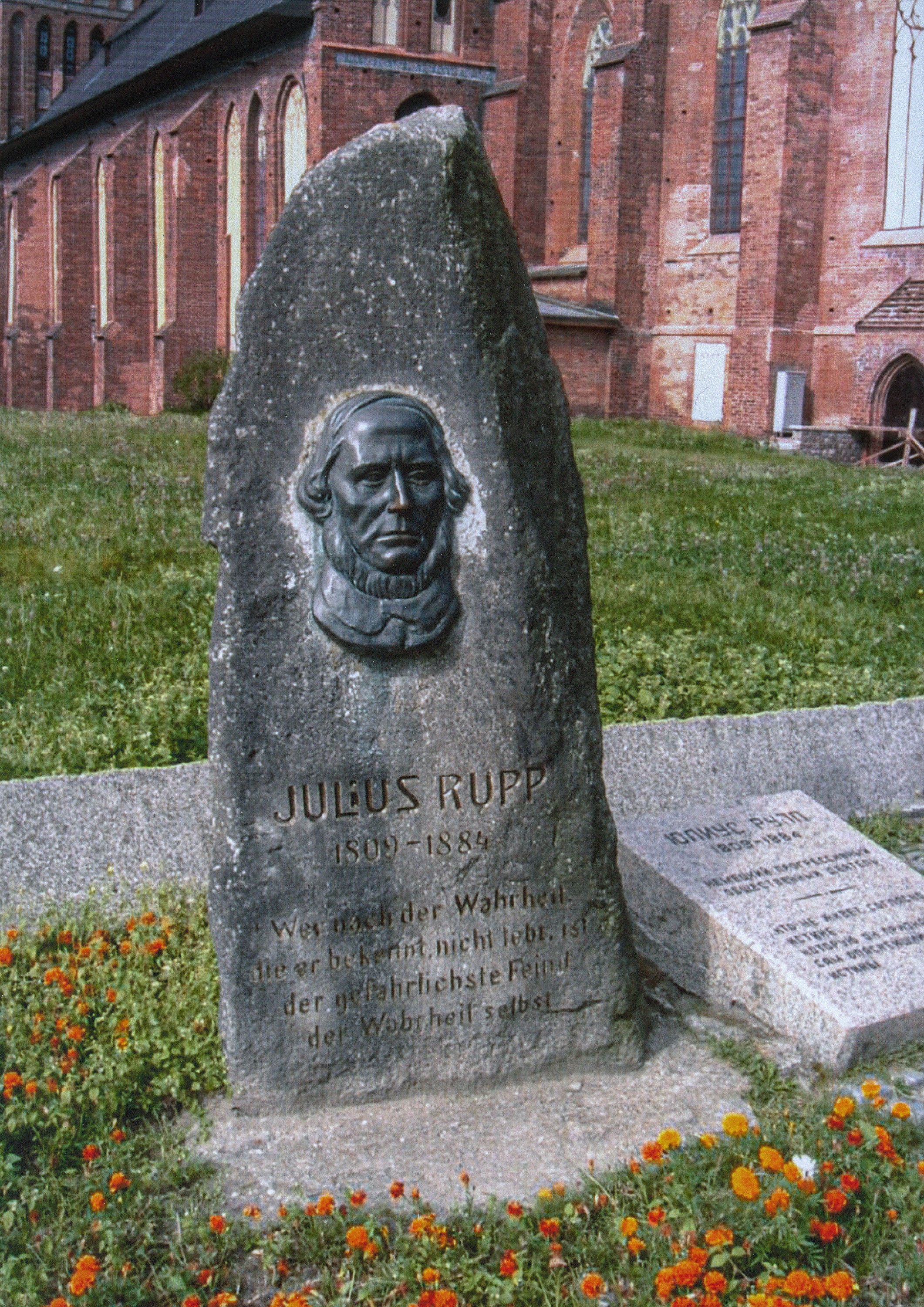 Das Bild zeigt einen großen, hochkant stehenden Gedenkstein aus grauem Naturstein, auf dem ein bronzenes Porträtrelief des Theologen Julius Rupp angebracht ist. Unter dem Relief sind sein Name, seine Lebensdaten (1809–1884) und ein Zitat eingraviert, das zur Wahrhaftigkeit mahnt. Der Stein steht auf einer Wiese vor der backsteinernen Fassade des Königsberger Doms, umgeben von kleinen orangefarbenen Blumen.