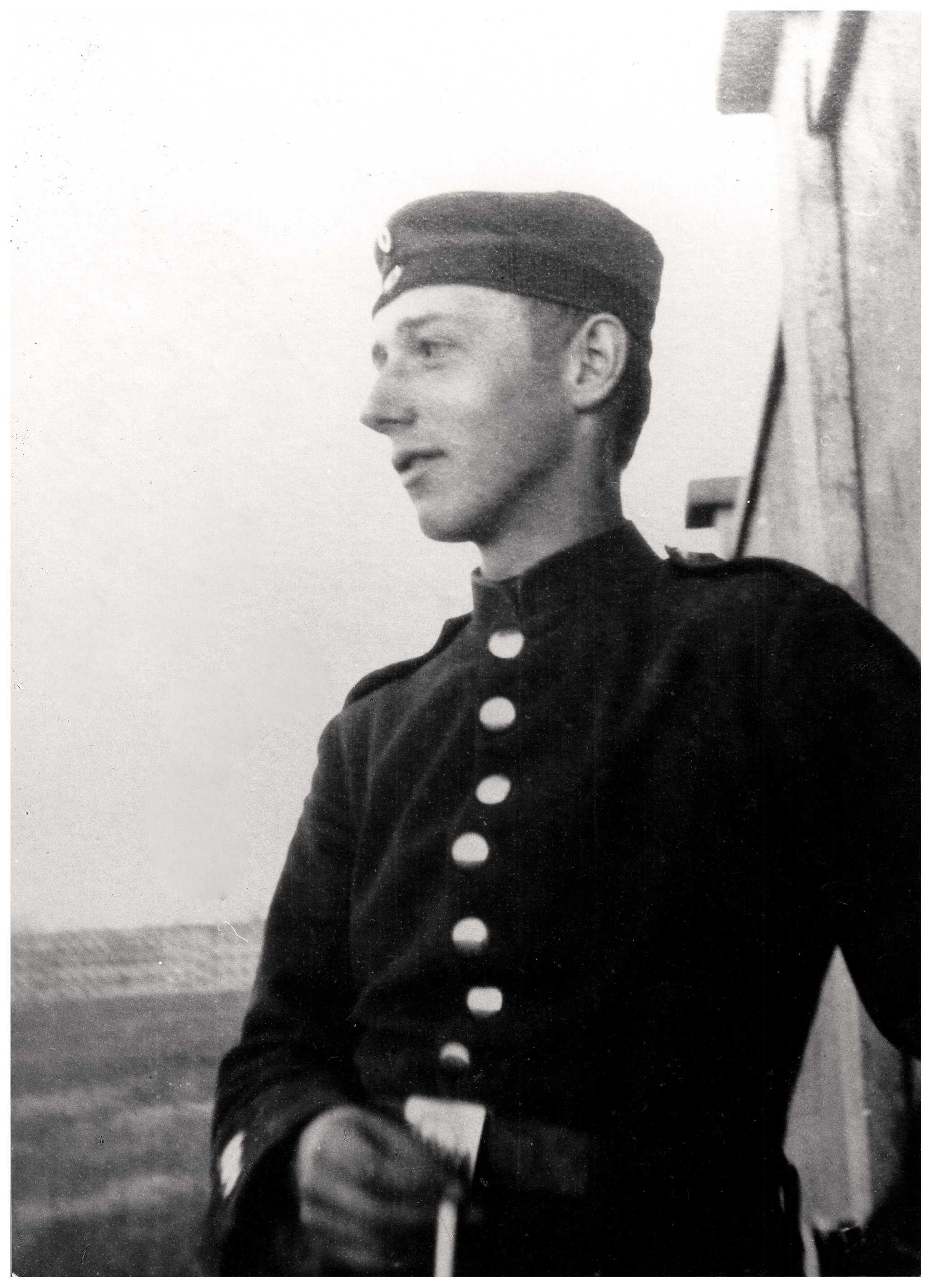 Dieses Schwarz-Weiß-Foto zeigt das Profil von Peter Kollwitz als jungen Mann in einer Militäruniform, der nach links blickt. Er trägt eine dunkle Jacke mit einer vertikalen Reihe heller Knöpfe und eine flache Feldmütze mit einem kleinen runden Abzeichen an der Vorderseite. Sein Gesichtsausdruck wirkt ruhig und nachdenklich, während er sich mit einem Arm leicht gegen eine Wand im Hintergrund lehnt.