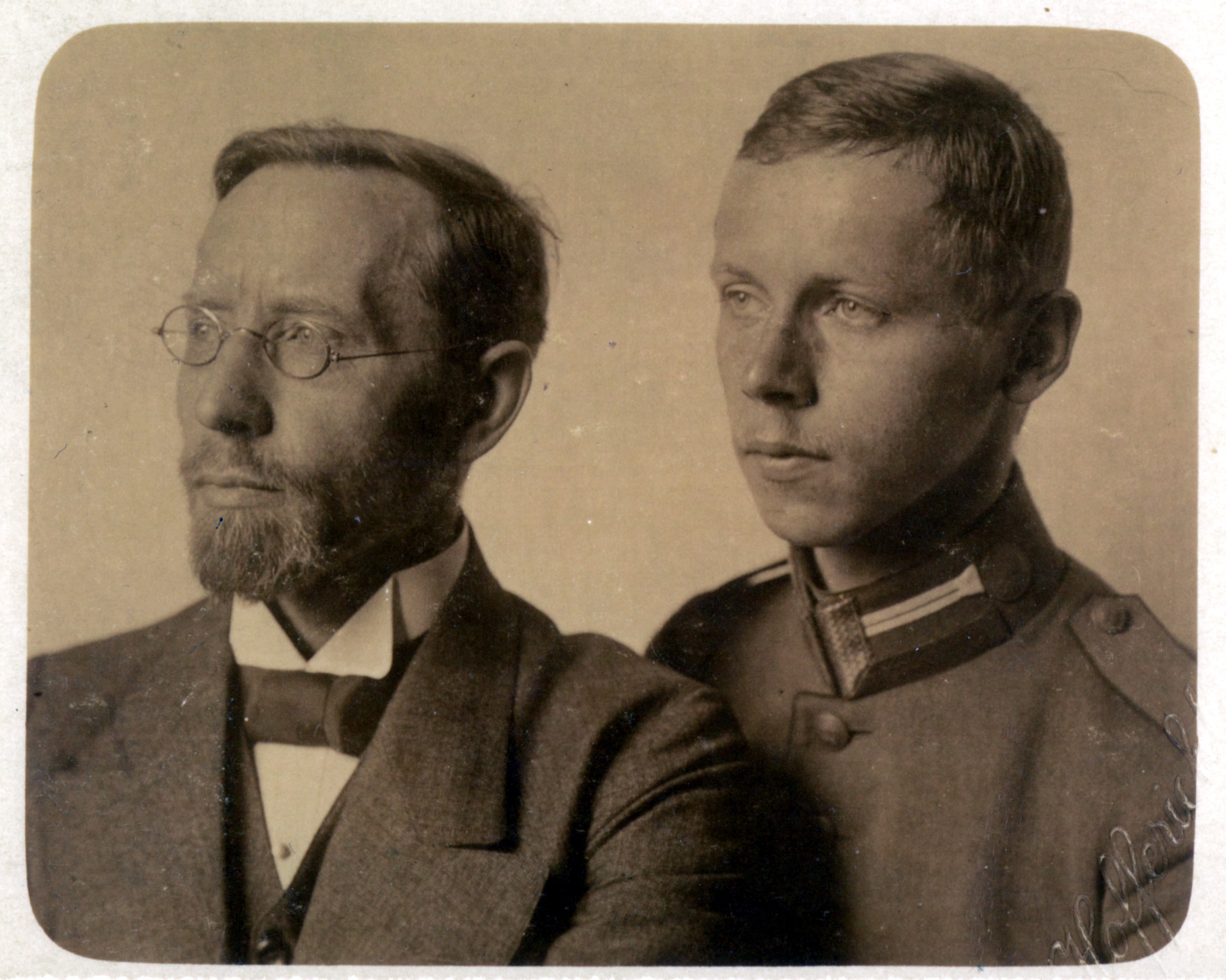 Das Schwarz-Weiß-Foto zeigt zwei Männer in einer Porträtaufnahme. Der ältere Mann im Vordergrund trägt eine Brille, einen Bart und formelle Kleidung mit Fliege, während der jüngere Mann dahinter eine Uniform trägt. Beide blicken ernst zur Seite.