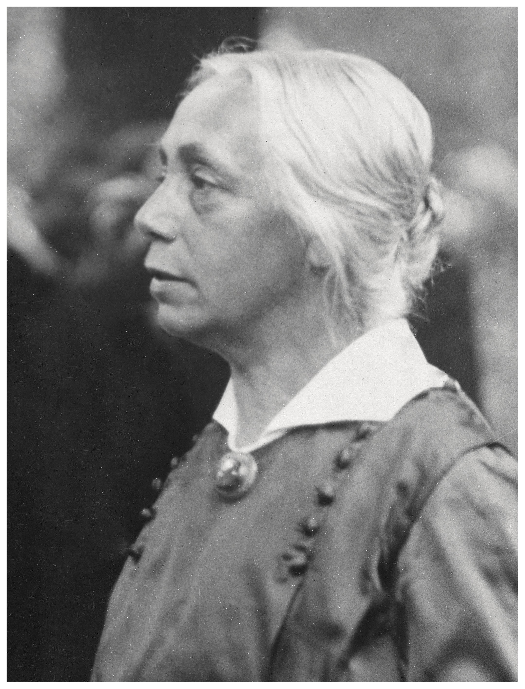 Das Schwarz-Weiß-Foto von Robert Sennecke zeigt Käthe Kollwitz im Profil mit hellem Haar, das am Hinterkopf zu einem ordentlichen Knoten gesteckt ist. Sie trägt eine dunkle Bluse mit einem markanten weißen Kragen und einer runden Brosche, während ihr Blick ernst und nachdenklich in die Ferne gerichtet ist.