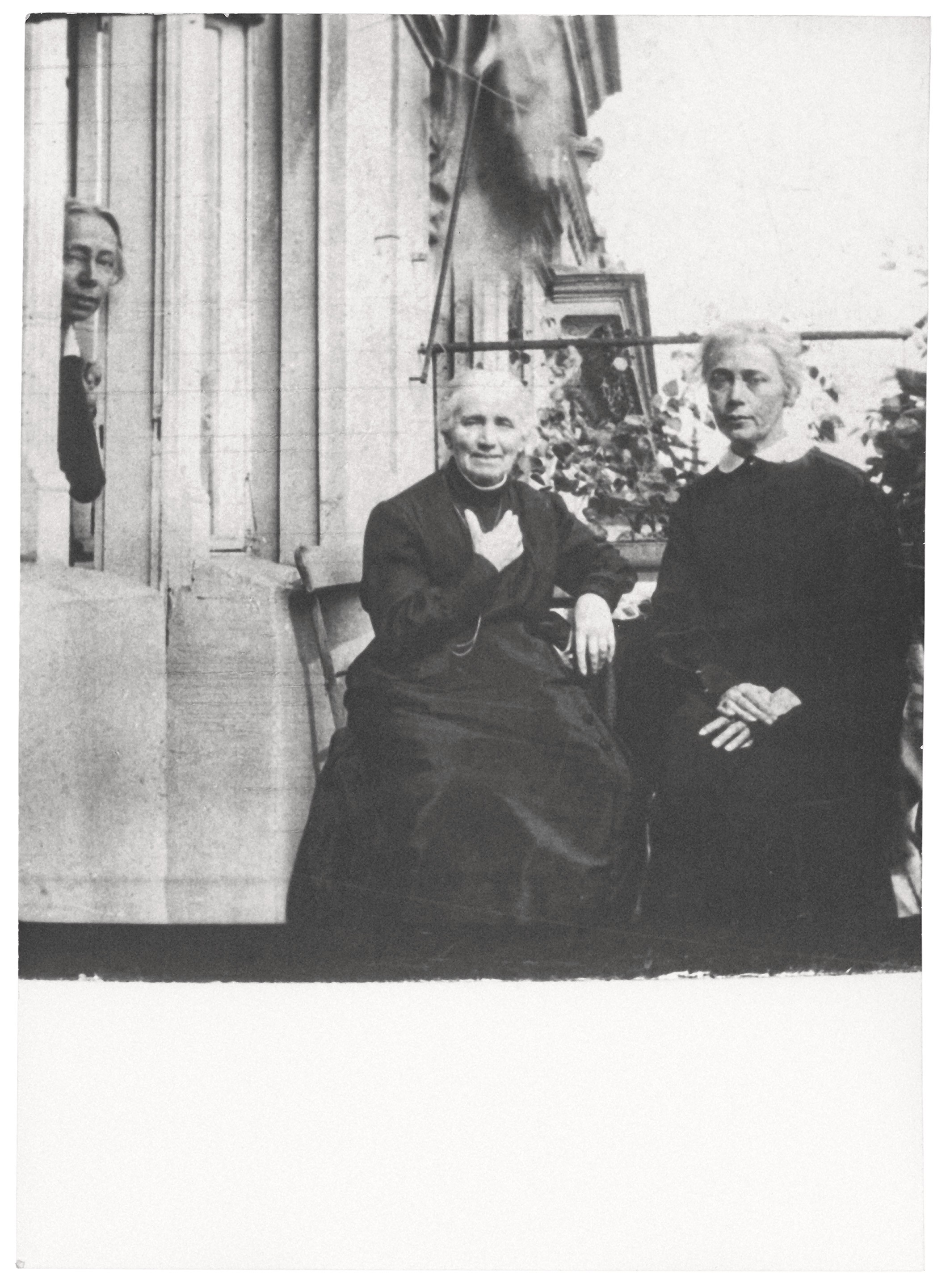 Das Schwarz-Weiß-Foto zeigt drei Frauen auf einem Balkon; im Zentrum sitzt eine ältere Frau, Katharina Schmidt, mit grauem Haar in einem dunklen Kleid, die ihre Hand sanft auf die Brust legt. Rechts daneben ihre Tochter Lise mit strengem Blick und weißem Kragen, während links im Hintergrund ihre Tochter Käthe aus einm Fenster hervorlugt. Die Szene wirkt ruhig und familiär, umgeben von steinernen Säulen und einigen Pflanzen im Hintergrund.