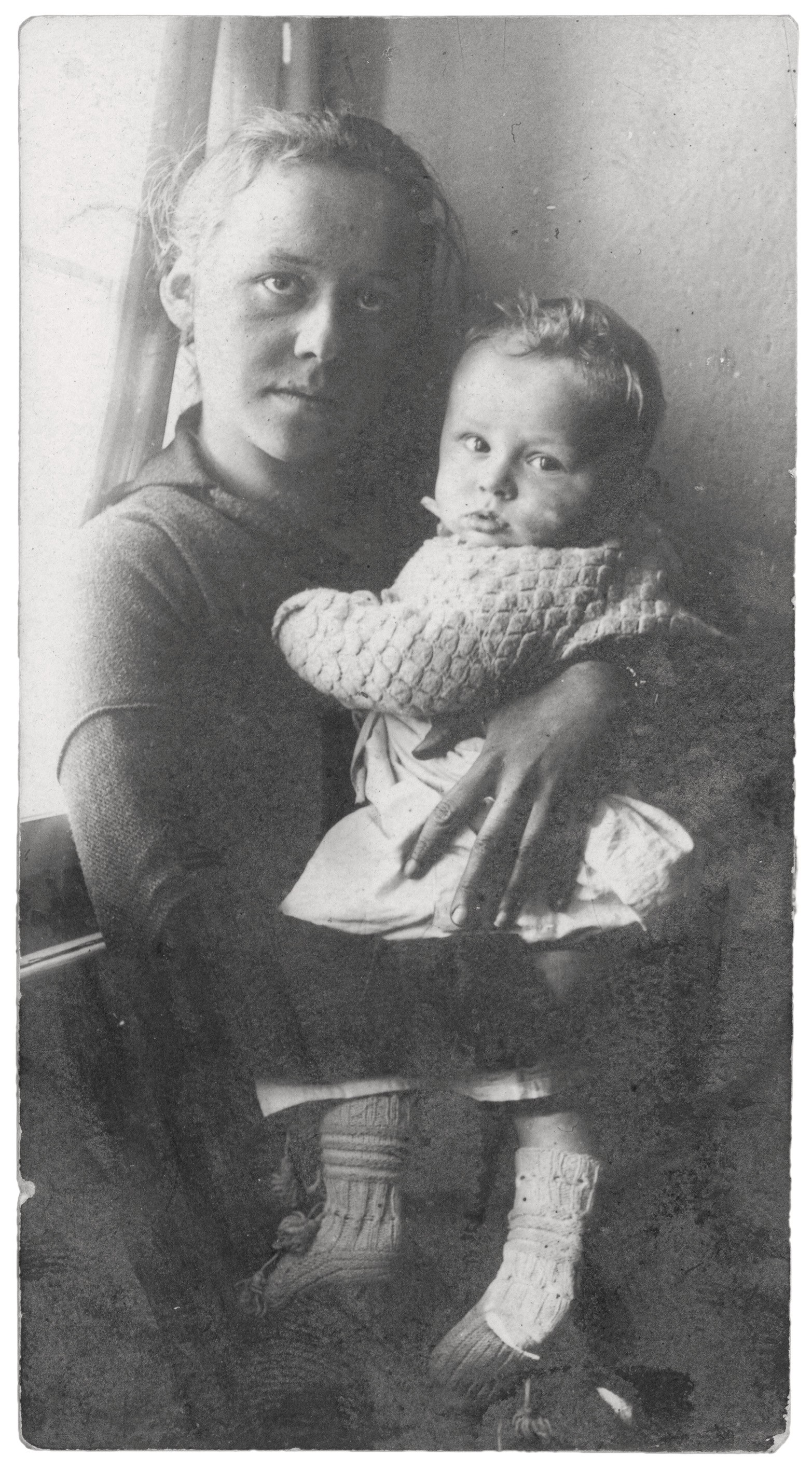 Eine Schwarz-Weiß-Fotografie zeigt Ottilie Ehlers-Kollwitz als junge Frau, die ihren kleinen Sohn Peter auf dem Schoß hält. Beide blicken ernst und direkt in die Kamera. Die Frau trägt ein dunkles Oberteil, während das Kind in eine helle, grob gestrickte Jacke und Söckchen gekleidet ist.