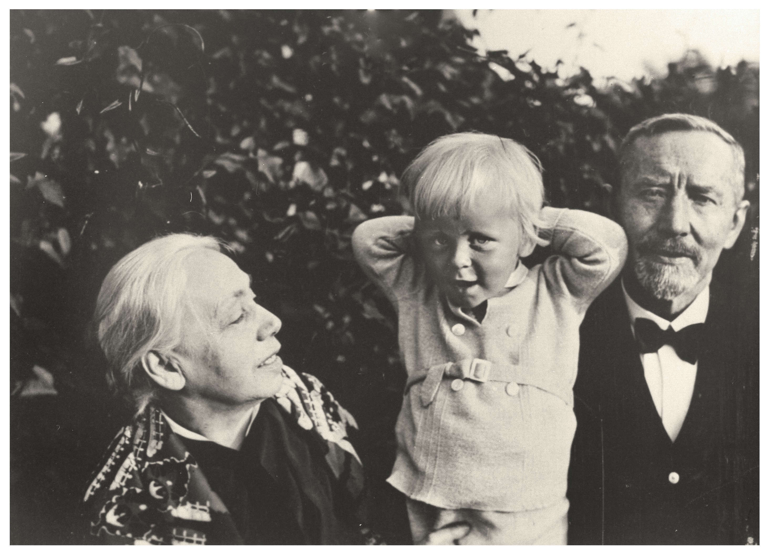 Das Schwarz-Weiß-Foto zeigt Käthe Kollwitz (links) und ihrem Mann Karl (rechts) sowie ihren Enkel Arnhe (mitte) in einem Garten, im Hintergrund dichtes Gebüsch. Käthe Kollwitz blickt lächelnd zu dem blonden Kind auf, das beide Hände hinter den Kopf hält, während rechts daneben Karl, ein bärtiger Mann im dunklen Anzug mit Fliege, direkt in die Kamera schaut. Die Szene wirkt wie ein privater, vertrauter Moment eines Familienporträts.