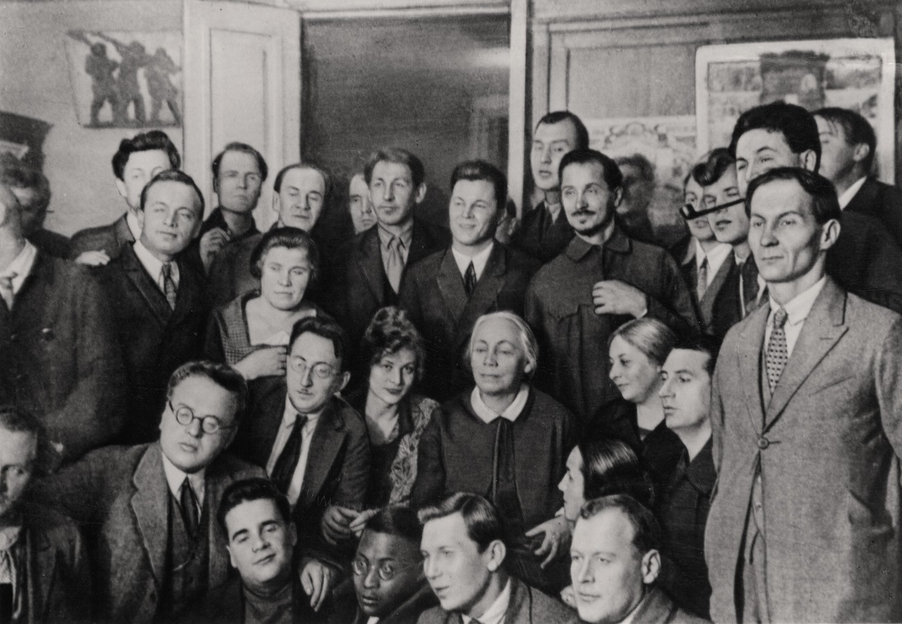 Dieses Schwarz-Weiß-Foto aus dem Jahr 1927 zeigt eine große Gruppe von etwa 25 Personen, überwiegend Männer in formellen Anzügen, die eng beieinander für ein Porträt posieren. Im Zentrum der Menge sitzt Käthe Kollwitz mit hellem Haar, gekleidet mit einem dunklem Kleid und weißem Kragen, während im Hintergrund an der Wand Skizzen und Plakate hängen. Die ernsten Gesichtsausdrücke und die klassische Kleidung verleihen der Aufnahme eine förmliche, historische Atmosphäre.