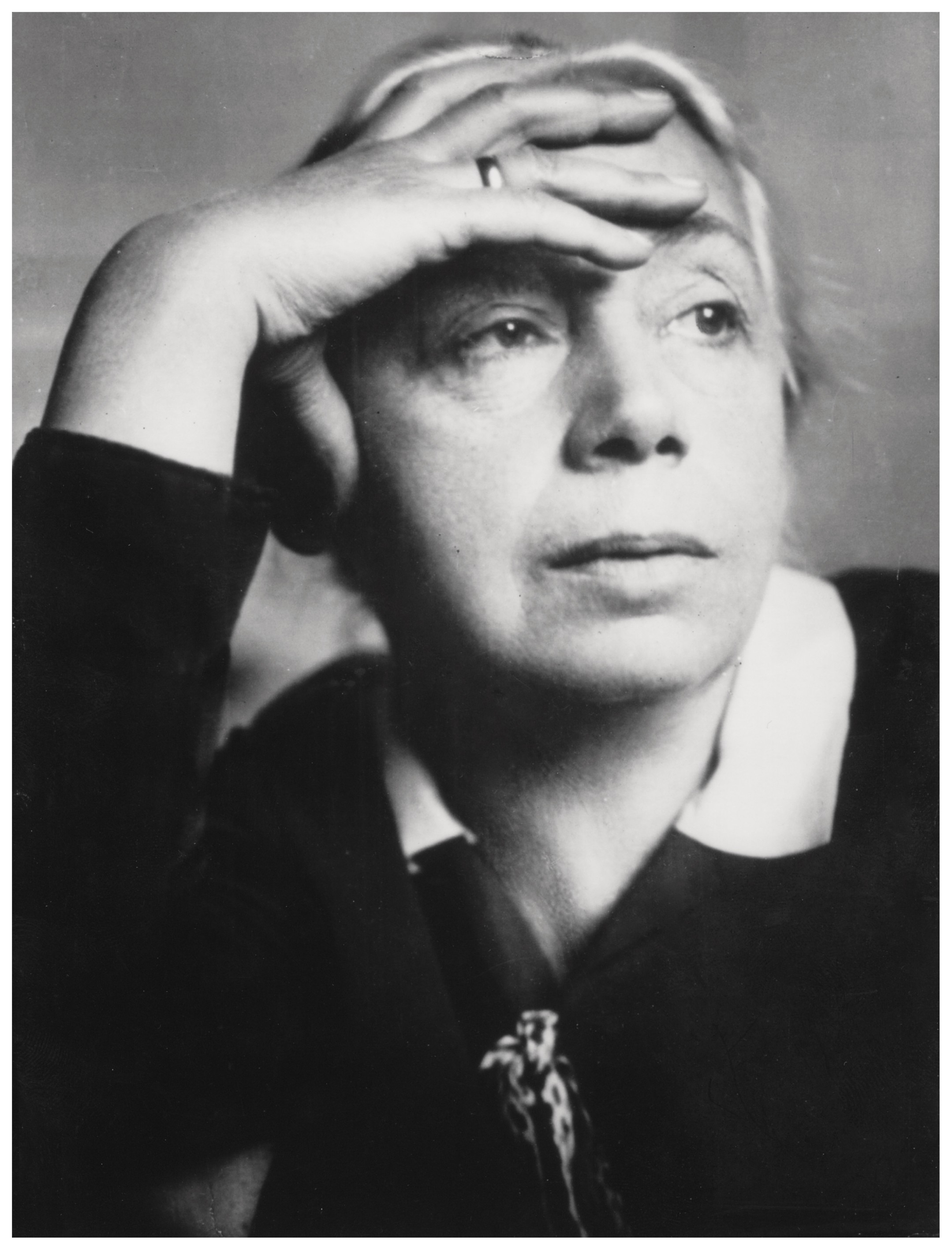 Dieses Schwarz-Weiß-Porträt von E.O. Hoppé aus dem Jahr 1928 zeigt die Künstlerin Käthe Kollwitz in einer nachdenklichen und ernsten Pose. Sie blickt am Betrachter vorbei in die Ferne, während ihre rechte Hand flach auf ihrer Stirn ruht, wobei ein schlichter Ring an ihrem Finger sichtbar ist. Ihr Gesichtsausdruck wirkt konzentriert und leicht melancholisch, was durch das Spiel von Licht und Schatten auf ihren Gesichtszügen unterstrichen wird.