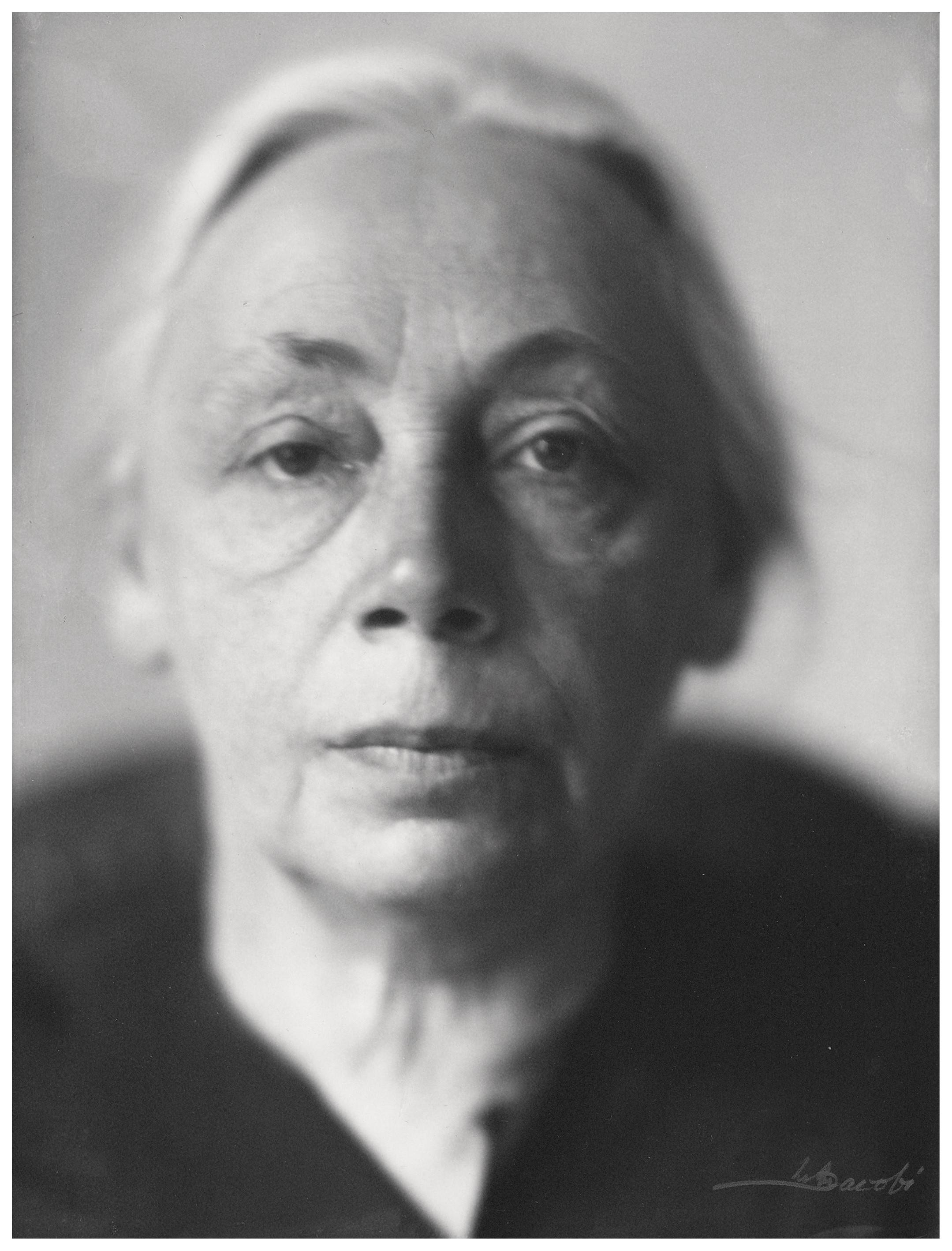 Diese Fotografie von Lotte Jacobi aus dem Jahr 1929 zeigt ein Schwarz-Weiß-Porträt der Künstlerin Käthe Kollwitz in einer Nahaufnahme, bei der ihr Gesicht fast das gesamte Format ausfüllt. Ihr Blick ist ruhig und direkt in die Kamera gerichtet, wobei ihr Ausdruck eine ernste, lebenserfahrene und zugleich würdevolle Tiefe vermittelt. Das Licht fällt seitlich ein, wodurch die feinen Linien ihres Gesichts betont werden und ein starker Kontrast zwischen den hellen Gesichtszügen und dem dunklen Hintergrund entsteht.