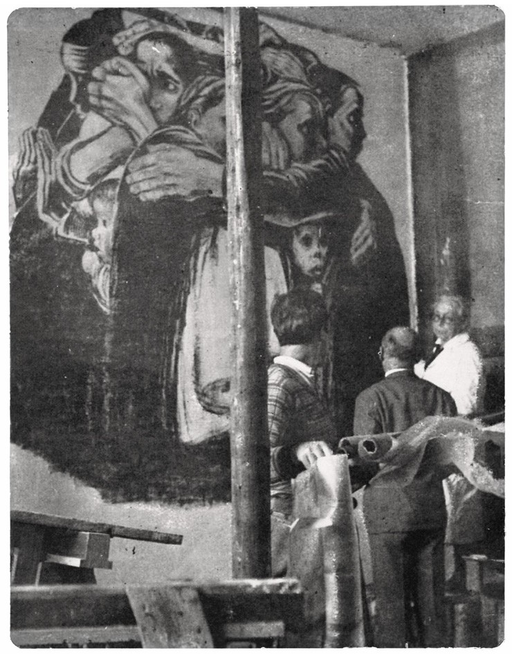 Dieses historische Schwarz-Weiß-Foto zeigt ein großes Wandgemälde an einer Innenwand, das eine Gruppe von Frauen darstellt, die sich eng aneinanderklammern und in ihrer Mitte kleine Kinder schützend umschließen. Im Vordergrund stehen Käthe Kollwitz, die Künstlerin, die den "Turm der Mütter" geschaffen hat, sowie zwei Männer in Alltagskleidung der 1920er oder 30er Jahre, die das monumentale Kunstwerk betrachten und gerade daran arbeiten.