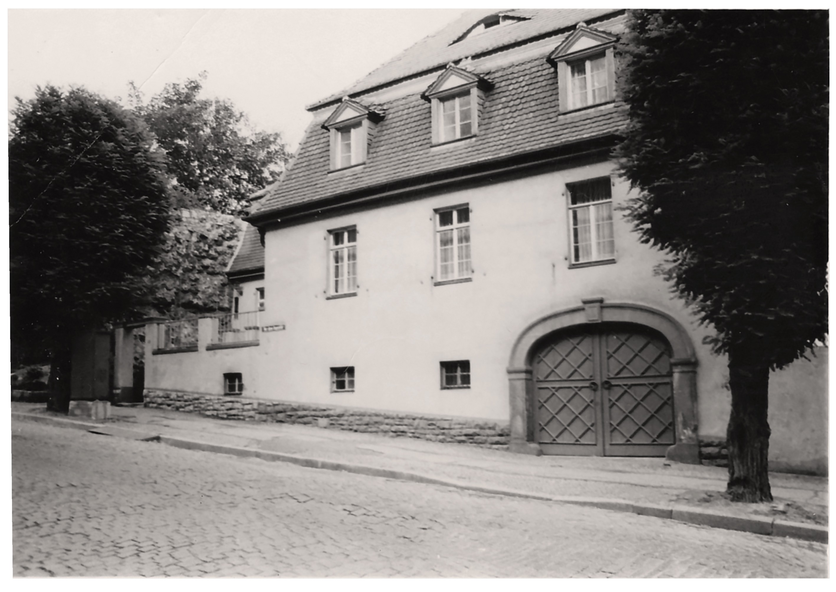 Das Bild zeigt ein zweistöckiges, helles Eckhaus in einer mit Kopfsteinpflaster gepflasterten Straße, das ein dunkles Mansarddach mit drei kleinen Dachgauben besitzt. Im Erdgeschoss befindet sich rechts ein großes, rundbogiges Holztor mit einem Rautenmuster, während links davon mehrere einfache Fenster in der Fassade angeordnet sind. Links und rechts wird das Gebäude von hohen, dicht belaubten Bäumen eingerahmt, die das Haus teilweise verschatten.