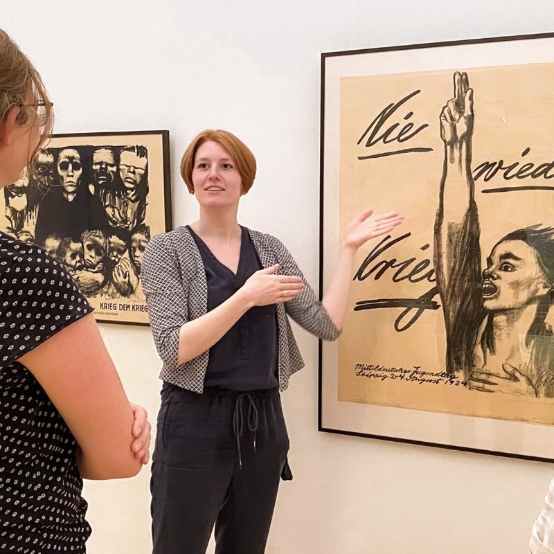 In hellen Ausstellungsräumen des Kollwitz Museum Köln erklärt eine Kunstvermittlerin mit ausladenden Handbewegungen zwei aufmerksam zuhörenden Besucherinnen das berühmte Antikriegsplakat 