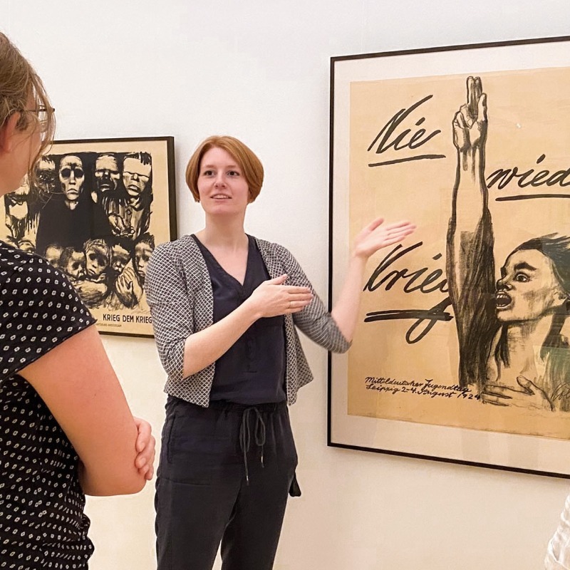 In hellen Ausstellungsräumen des Kollwitz Museum Köln erklärt eine Kunstvermittlerin mit ausladenden Handbewegungen zwei aufmerksam zuhörenden Besucherinnen das berühmte Antikriegsplakat 