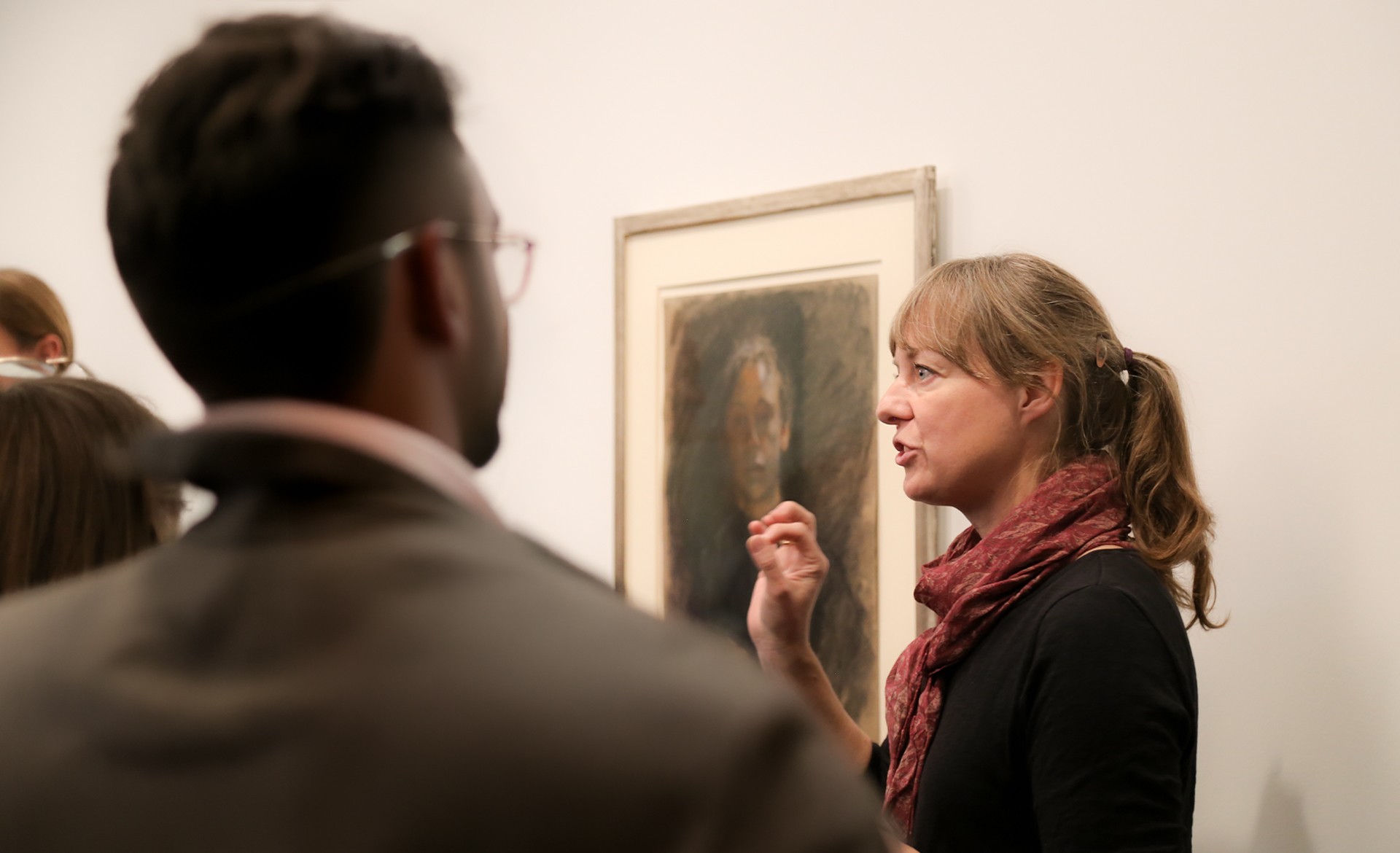 Eine Kunstvermittlerin mit langem Haar und Schal steht im Profil vor einem gerahmten Selbstporträt von Käthe Kollwitz und erklärt es gestikulierend einer Besuchergruppe, von der ein Mann im Vordergrund unscharf von hinten zu sehen ist. Das gezeigte Kunstwerk ist eine farbige Pastellzeichnung, die das Gesicht der Künstlerin in einer frontalen, nachdenklichen Ansicht zeigt.