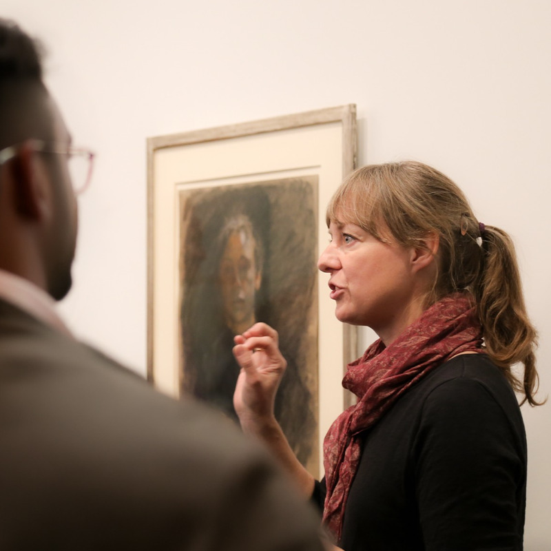 Eine Kunstvermittlerin mit langem Haar und Schal steht im Profil vor einem gerahmten Selbstporträt von Käthe Kollwitz und erklärt es gestikulierend einer Besuchergruppe, von der ein Mann im Vordergrund unscharf von hinten zu sehen ist. Das gezeigte Kunstwerk ist eine farbige Pastellzeichnung, die das Gesicht der Künstlerin in einer frontalen, nachdenklichen Ansicht zeigt.