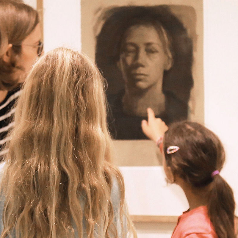 Das Bild zeigt eine Frau und drei junge Mädchen, die in einem Museum dicht vor einem großen, gerahmten Porträt stehen. Während die Kunstvermittlerin und zwei der Kinder das Werk aufmerksam betrachten, deutet ein Mädchen mit dem Finger direkt auf das Gesicht des Selbstbildnisses von Käthe Kollwitz.