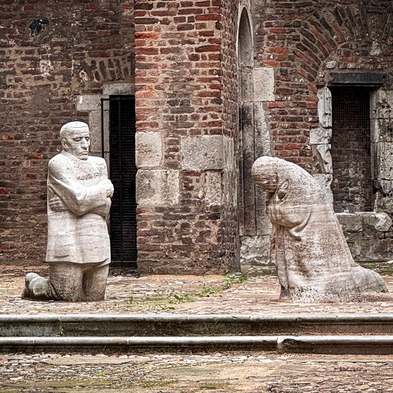 Das Foto von Christian Nitz zeigt das Mahnmal »Trauernde Eltern« von Käthe Kollwitz, das im offenen Innenraum der Kirchenruine Alt St. Alban in Köln aufgestellt ist. Inmitten der verwitterten Backsteinmauern knien sich zwei lebensgroße, blockhaft massive Steingestalten in stillem Schmerz gegenüber: Links verharrt der Vater mit starr geradeaus gerichtetem Blick und eng vor der Brust verschränkten Armen, während rechts die Mutter tief gebeugt und in sich gekehrt um den gemeinsamen Sohn trauert.