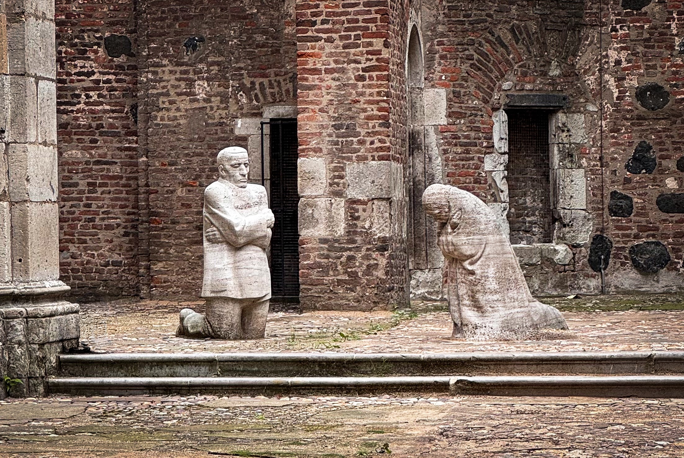 Das Foto von Christian Nitz zeigt das Mahnmal »Trauernde Eltern« von Käthe Kollwitz, das im offenen Innenraum der Kirchenruine Alt St. Alban in Köln aufgestellt ist. Inmitten der verwitterten Backsteinmauern knien sich zwei lebensgroße, blockhaft massive Steingestalten in stillem Schmerz gegenüber: Links verharrt der Vater mit starr geradeaus gerichtetem Blick und eng vor der Brust verschränkten Armen, während rechts die Mutter tief gebeugt und in sich gekehrt um den gemeinsamen Sohn trauert.