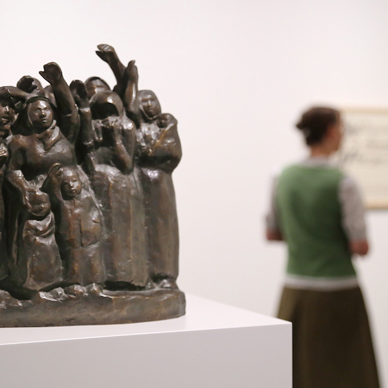 Das Bild zeigt die dunkle Bronzeplastik einer Gruppe eng beieinanderstehender, winkender Frauen und Kinder auf einem weißen Sockel, während im unscharfen Hintergrund eine Museumsbesucherin ein gerahmtes Plakat von Käthe Kollwitz betrachtet..