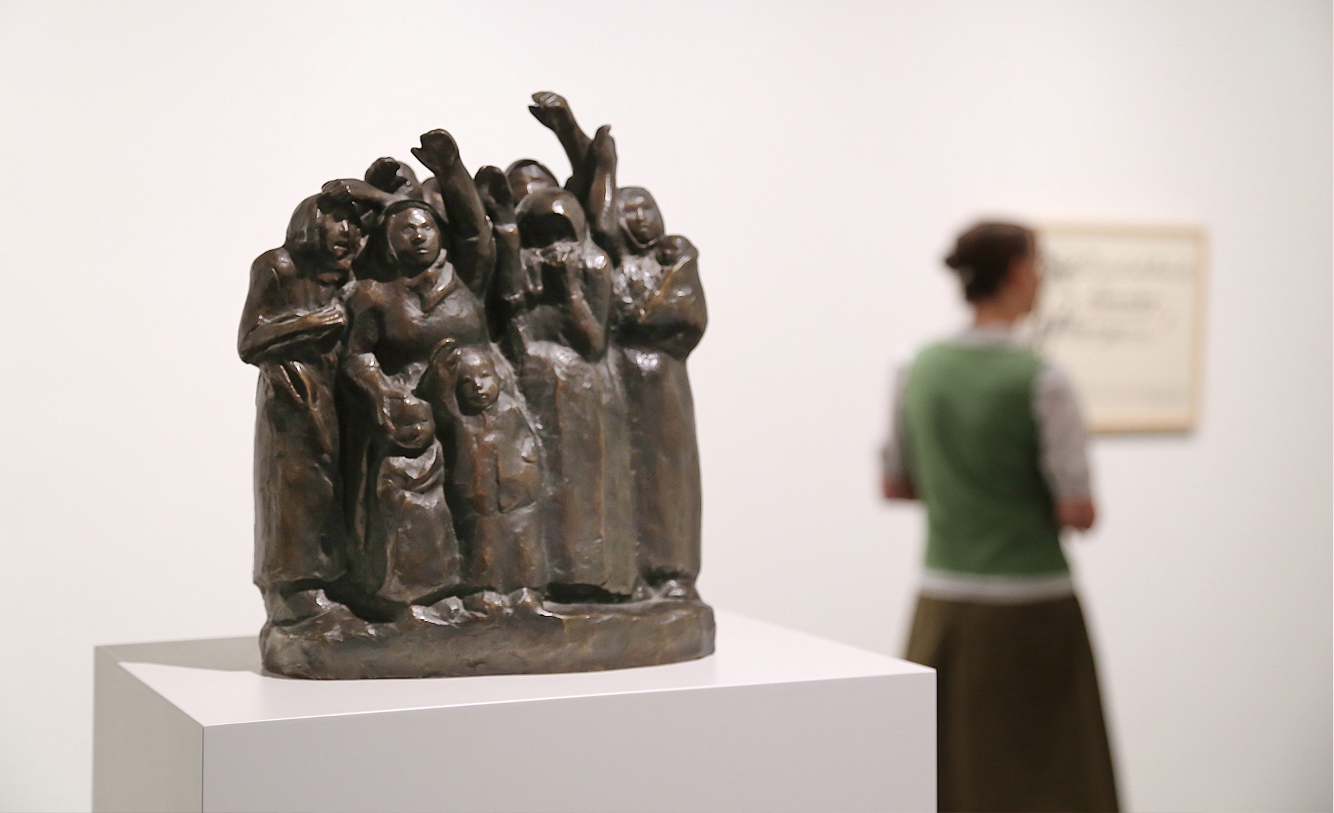 Das Bild zeigt die dunkle Bronzeplastik einer Gruppe eng beieinanderstehender, winkender Frauen und Kinder auf einem weißen Sockel, während im unscharfen Hintergrund eine Museumsbesucherin ein gerahmtes Plakat von Käthe Kollwitz betrachtet.. 