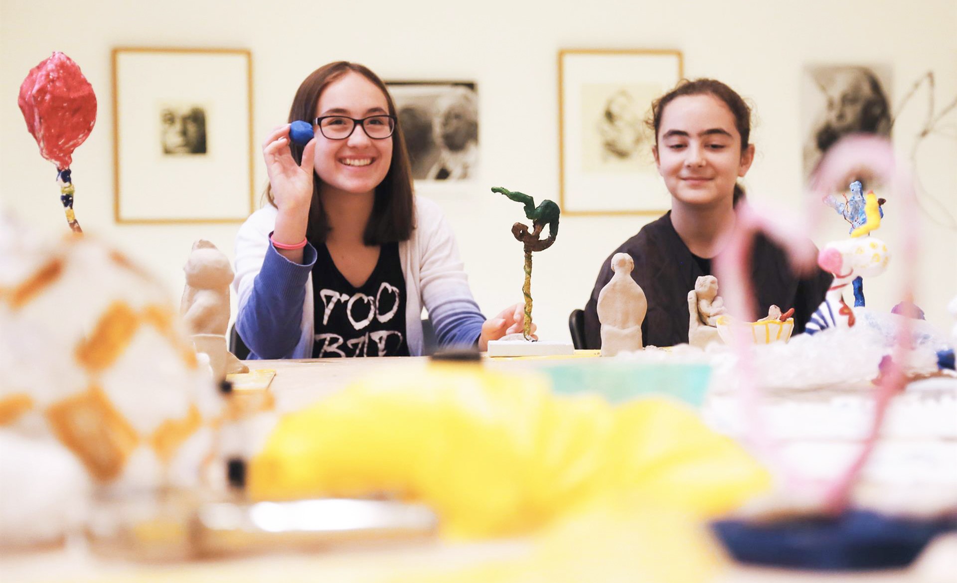 Zwei junge Mädchen sitzen während eines Workshops lächelnd an einem Werktisch in den hellen Räumen des Kollwitz Museum Köln und präsentieren stolz ihre kleinen, handgefertigten Skulpturen. Im unscharfen Vordergrund liegen bunte Bastelmaterialien und Werkzeuge, während im Hintergrund gerahmte Kunstwerke von Käthe Kollwitz an den weißen Wänden hängen.