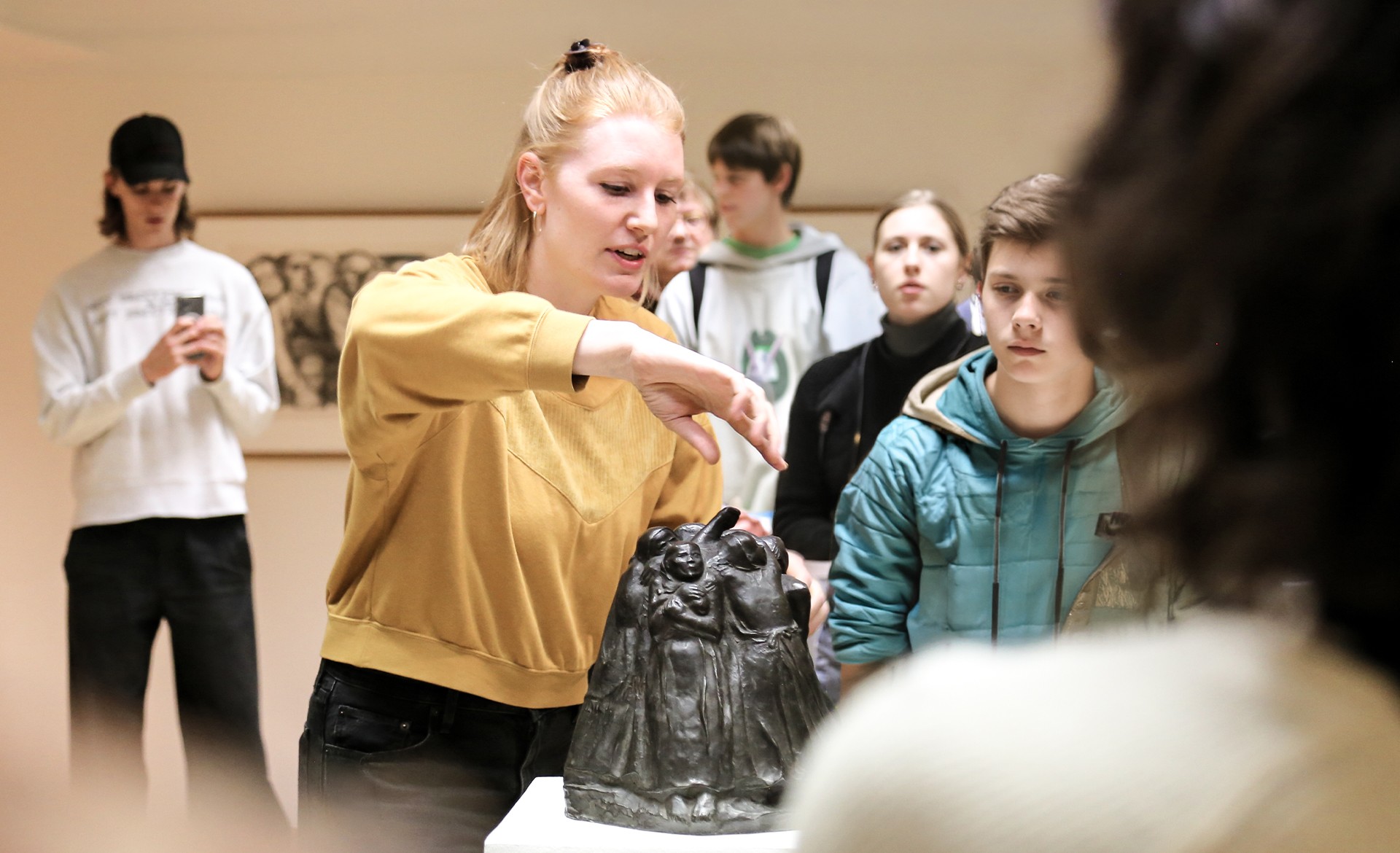 Ein Foto zeigt eine Kunstvermittlerin im Kollwitz Museum Köln. Sie trägt ein senfgelbes Sweatshirt neigt sich erklärend über die kleine, dunkle Bronzeplastik, den  „Turm der Mütter“ von Käthe Kollwitz, die auf einem weißen Sockel steht. Sie ist umringt von einer Schülergruppe , die aufmerksam die kreisförmig angeordneten, schützenden Frauenfiguren der Skulptur betrachten und den Ausführungen zuhören.