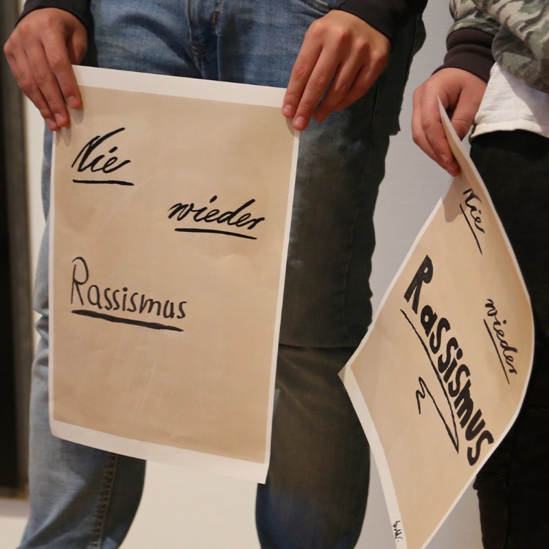 Drei Jugendliche halten selbst gestaltete Plakate mit der Aufschrift „Nie wieder Rassismus“ in die Kamera, wobei die Schriftart des Wortes „Nie“ als Hommage an das berühmte Plakat von Käthe Kollwitz gestaltet ist. Die Schilder entstanden während eines kreativen Workshops im Kollwitz Museum Köln, bei dem sich die Teilnehmer künstlerisch mit aktuellen gesellschaftlichen Themen auseinandersetzen.