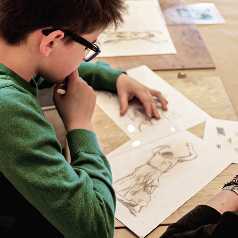 Das Bild zeigt einen Jungen im Profil und die Schulter eines Mädchens, die gemeinsam an einem Tisch über Plexiglasscheiben gebeugt sind. Konzentriert ritzen sie mit feinen Werkzeugen eine Szene aus Käthe Kollwitz' Zyklus „Bauernkrieg“ in das Material, um eine Druckplatte für einen Tiefdruck zu erstellen.