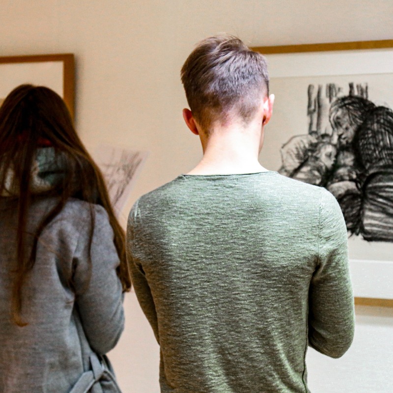 Zwei Jugendliche stehen im Kollwitz Museum Köln vor gerahmten Werken von Käthe Kollwitz und skizzieren konzentriert deren Motive auf eigene Blätter. Der junge Mann im Vordergrund betrachtet eine ausdrucksstarke Kreidezeichnung einer kauernden Mutter mit Kind, ein Mädchen neben ihm mit langem braunen Haar zeichnet ebenfalls.