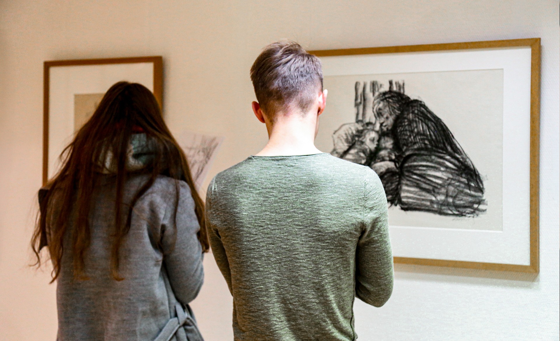 Zwei Jugendliche stehen im Kollwitz Museum Köln vor gerahmten Werken von Käthe Kollwitz und skizzieren konzentriert deren Motive auf eigene Blätter. Der junge Mann im Vordergrund betrachtet eine ausdrucksstarke Kreidezeichnung einer kauernden Mutter mit Kind, ein Mädchen neben ihm mit langem braunen Haar zeichnet ebenfalls.