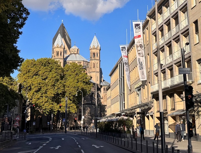 The photograph shows the modern, light-coloured façade of the Käthe Kollwitz Museum on Cologne’s bustling Neumarkt, from which long vertical banners featuring the theme of the current special exhibition hang. In the background, the striking grey towers of the Romanesque Basilica of St Apostles rise into the blue sky behind the green treetops.