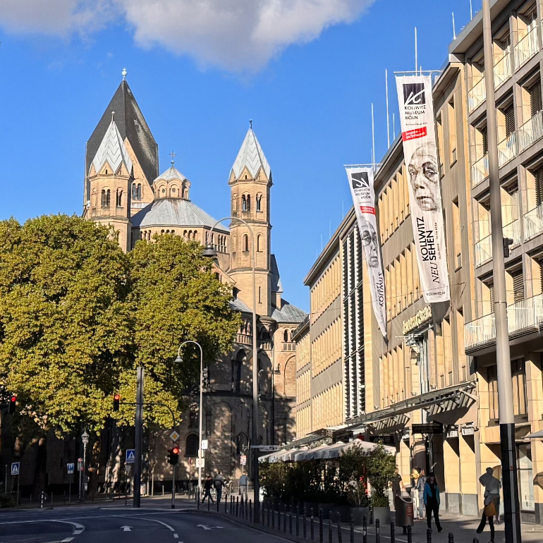 The photograph shows the modern, light-coloured façade of the Käthe Kollwitz Museum on Cologne’s bustling Neumarkt, from which long vertical banners featuring the theme of the current special exhibition hang. In the background, the striking grey towers of the Romanesque Basilica of St Apostles rise into the blue sky behind the green treetops.