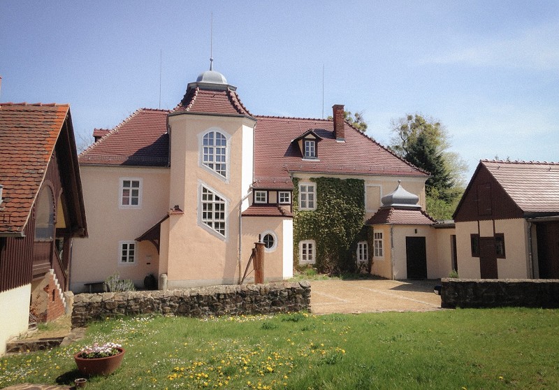The picture shows the Rüdenhof in Moritzburg – the place where Käthe Kollwitz died. It is a light-coloured, multi-storey building with a striking hexagonal stair tower and reddish tiled roofs, surrounded by a low natural stone wall. In the foreground lies a green meadow dotted with yellow dandelion flowers and a small planter, whilst the main building is partly covered in ivy and flanked by smaller outbuildings.