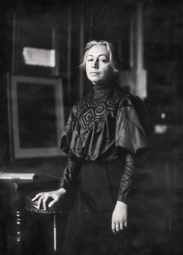 A photograph of Käthe Kollwitz from 1906, standing, with her 1901 etching »Carmagnole« in the background. The 39-year-old looks directly at the viewer, resting her right hand on a stool-like piece of furniture; she wears an elegant dark, high-necked blouse with striking embroidery on the chest and voluminous sleeves, which are puffed at the shoulder and taper into close-fitting cuffs at the wrist. The photographer is Philipp Kester. A reproduction print from the artist’s estate is held in the archives of the Kollwitz Museum Köln, Inv. No. F-01.a-06.