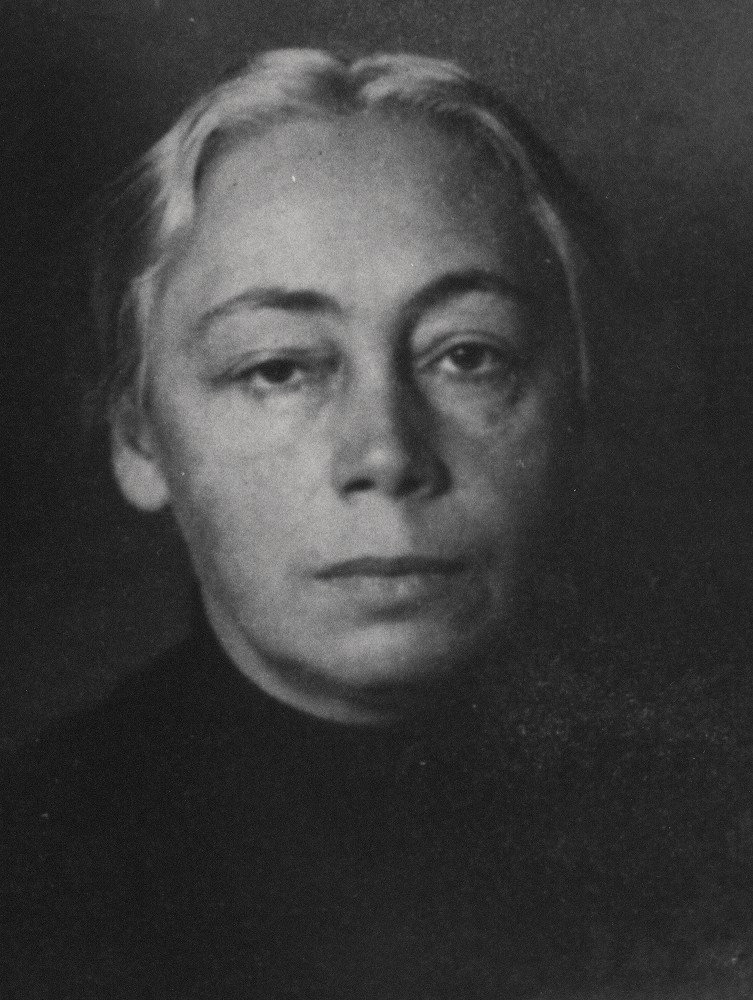 A black-and-white photograph shows Käthe Kollwitz’s face in a close-up frontal shot against a dark, plain background. It was taken around 1914–15, following the death of her son Peter. Photographer unknown. Her light grey hair is combed back strictly with a centre parting, emphasising her high forehead and distinct facial features. She looks directly into the camera with a serious, world-weary expression. Her gaze appears tired and dejected. 
This image, a detail from a portrait with her son Hans, is part of the artist’s estate in the archives of the Kollwitz Museum Köln, Inv.: No. F 01.b-07.