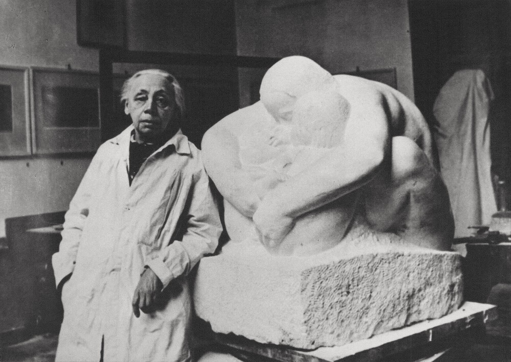 This black-and-white photograph, believed to have been taken in 1938, shows Käthe Kollwitz in her studio on Klosterstraße in Berlin. The artist, aged around 71, stands to the left of the plaster cast of her sculpture »Mother with Two Children«. She is wearing a long, light-coloured smock and gazes directly into the camera with a serious expression, whilst her left hand rests loosely on the plinth of her work. The larger-than-life sculpture depicts a mother figure bent protectively over two small children, holding them tightly in her arms. In the background, the plain walls of the studio can be seen, featuring a few framed pictures and other covered objects, which emphasises the working atmosphere. Photographer unknown. A reproduction print of this photograph from the artist’s estate is held in the archives of the Kollwitz Museum Köln, Inv.: No. F 01.a-37.