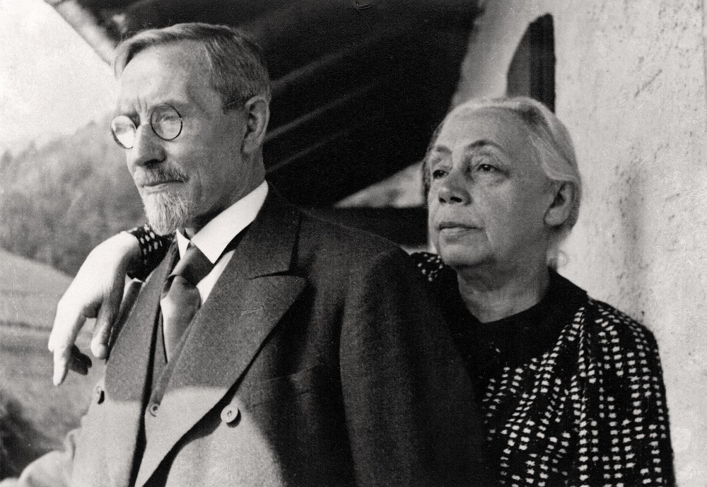 This black-and-white photograph from 1935 shows Käthe and Karl Kollwitz in a half-length portrait, taken outdoors in front of their holiday home during their holiday in Bad Reichenhall. The elegantly dressed Karl Kollwitz stands in the foreground, wearing glasses, a tie and a dark coat, whilst gazing thoughtfully into the distance. Käthe Kollwitz stands slightly behind him. She is wearing a dark suit with a white pattern and has placed her arm familiarly on his shoulder. She looks in the same direction with a serious, composed expression. Photographer unknown. A reproduction print of this photograph from the artist’s estate is held in the archives of the Kollwitz Museum Köln, Inv.: No. F 01.b-31.