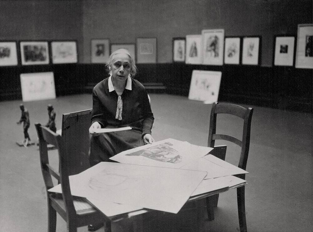 The black-and-white photograph from 1927 shows Käthe Kollwitz in an exhibition hall at the Prussian Academy of Arts in Berlin. The 60-year-old is wearing a simple, dark suit with a white collar, beneath which a light-coloured ribbon is tied in a manner resembling a tie.  
She is sitting in front of a makeshift work surface consisting of two wooden chairs with high backs pushed together. The seats form a sort of table. On it lie some of her works, large sheets of paper, one of which she lifts slightly with both hands to select it for the planned exhibition. In the background, on the walls of the spacious room, numerous framed prints are already hung in a row. Her gaze is directed straight at the camera and appears serious and scrutinising. Photographer unknown. A reproduction print of this photograph from the artist’s estate is held in the archives of the Kollwitz Museum Köln, Inv.: No. F 01.e-01.