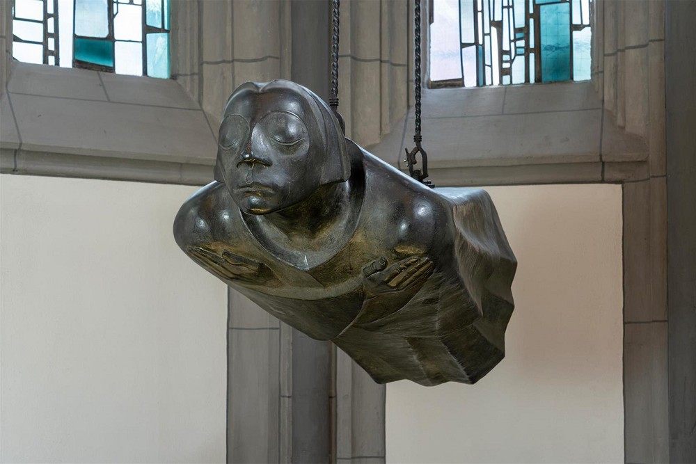 The photograph by Chris Franken shows the bronze sculpture »Der Schwebende« (The Floating Angel) by Ernst Barlach — the second cast after the original working model of 1939 — in its present location in the Antoniterkirche in Cologne, seen frontally and turned to the left. The life-size bronze figure, hovering horizontally, hangs freely in the air from two chains, its straight body and arms crossed over the chest radiating a deep serenity. The face of the sculpture has closed eyes and seems absorbed in itself, while the flowing form of the robe reinforces the impression of a gentle flight.