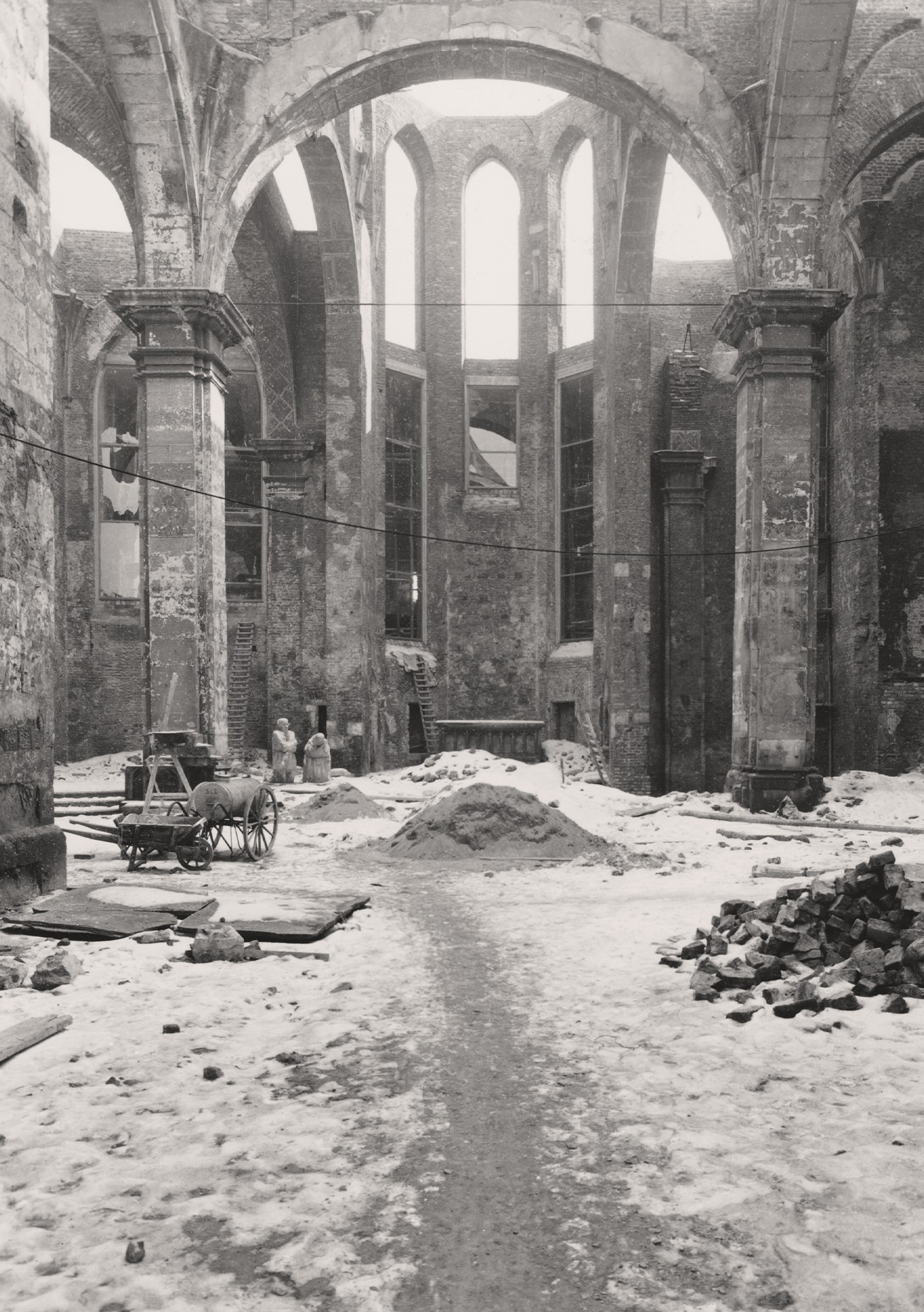 This black-and-white photograph shows a view inside the ruins of the Old St Alban’s Church in Cologne during the installation of the pair of sculptures ‘Grieving Parents’ in the winter months of 1955. The courtyard is covered in snow; in the foreground on the right is a pile of stones, on the left a cart with large wooden wheels and tools; in the background, the sculptures can be seen already in place.