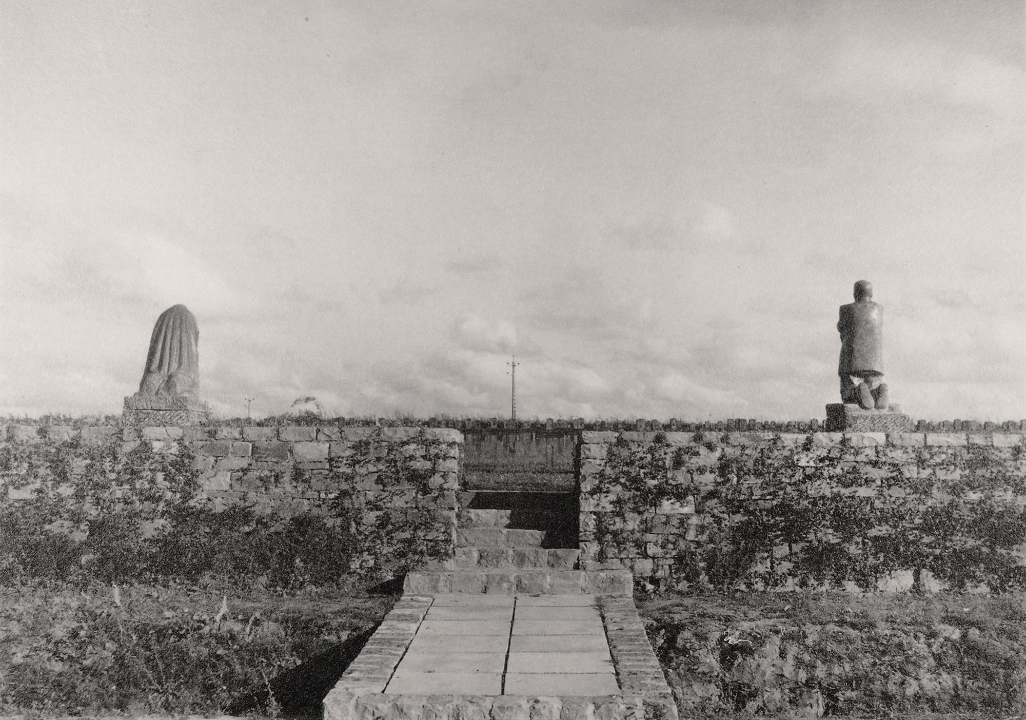 This black-and-white photograph from 1932 shows a view of the Roggeveld Military Cemetery from the outside: a narrow path paved with stone slabs leads through an opening in the low rubble-stone wall into the cemetery; behind it, to the right of the entrance, stands the sculpture of the mother, and to the left that of the father, both seen from behind.