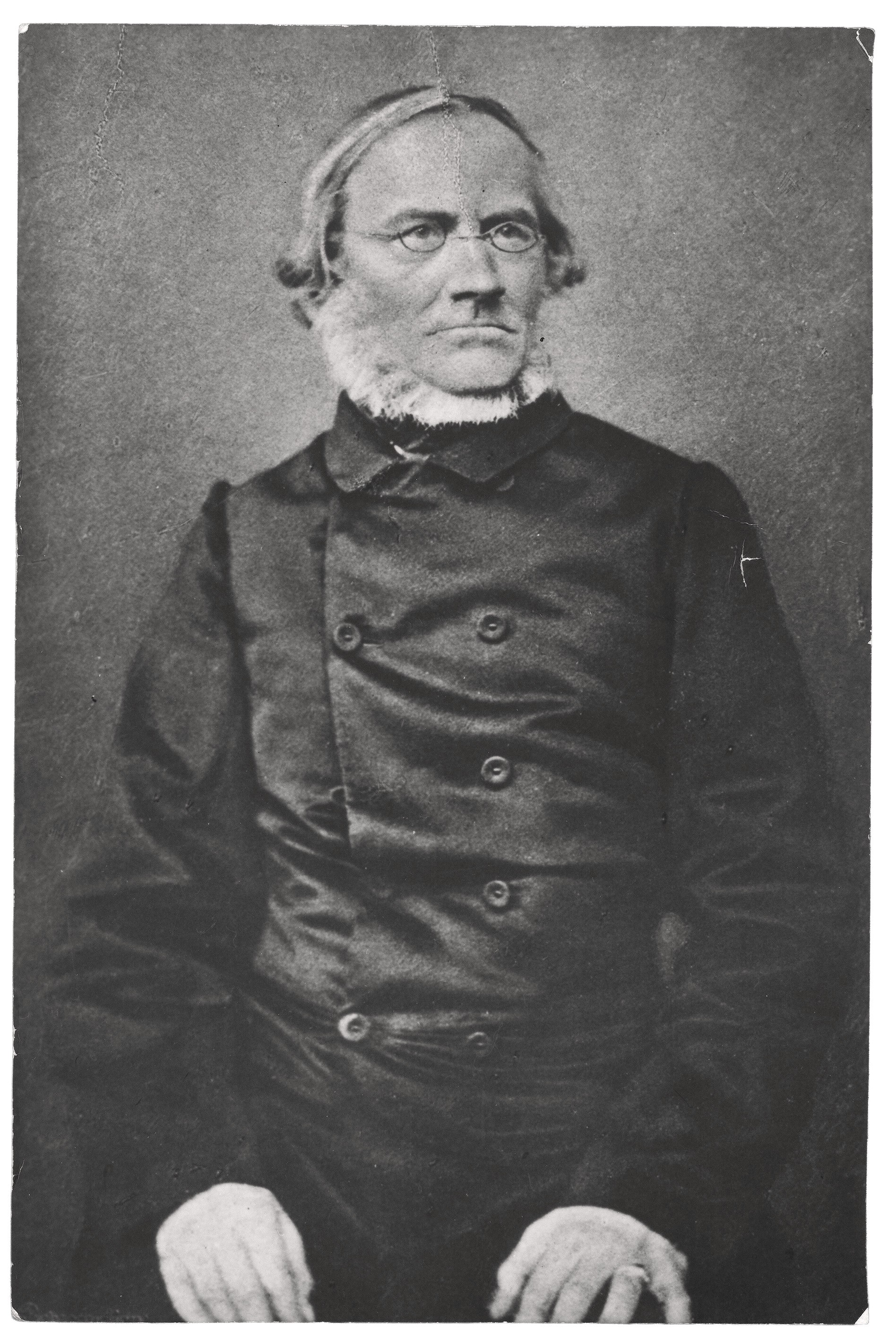 The black-and-white portrait depicts Julius Rupp (1809–1884) as an elderly man, shown with a serious gaze directed slightly to the side. He wears a dark, high-buttoned jacket with prominent buttons, a pair of narrow spectacles, and a distinctive white chin curtain beard that frames his face beneath the chin. His hands rest calmly on his knees in the foreground of the image.