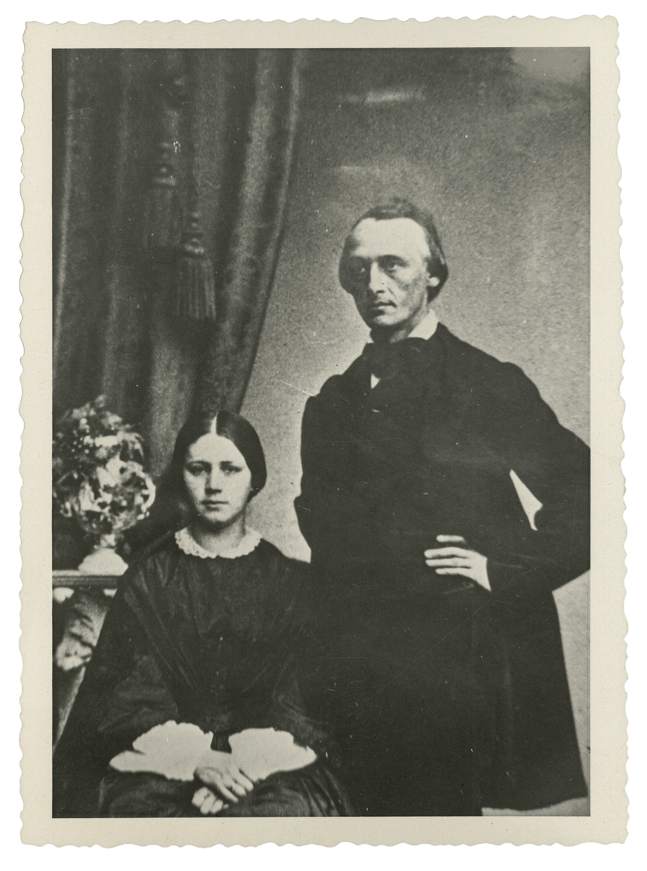 The image shows a black-and-white portrait of Carl Schmidt (1825–1898), standing, and his wife Katharina Schmidt, née Rupp (1837–1925), seated to his left, dressed in formal mid-nineteenth-century attire. Carl Schmidt wears a dark frock coat and rests one hand on his hip, while his wife, wearing a dark dress with a light lace collar and cuffs, has her hands folded in her lap. In the background, a heavy drape with a tassel and a small table with a floral arrangement can be seen.