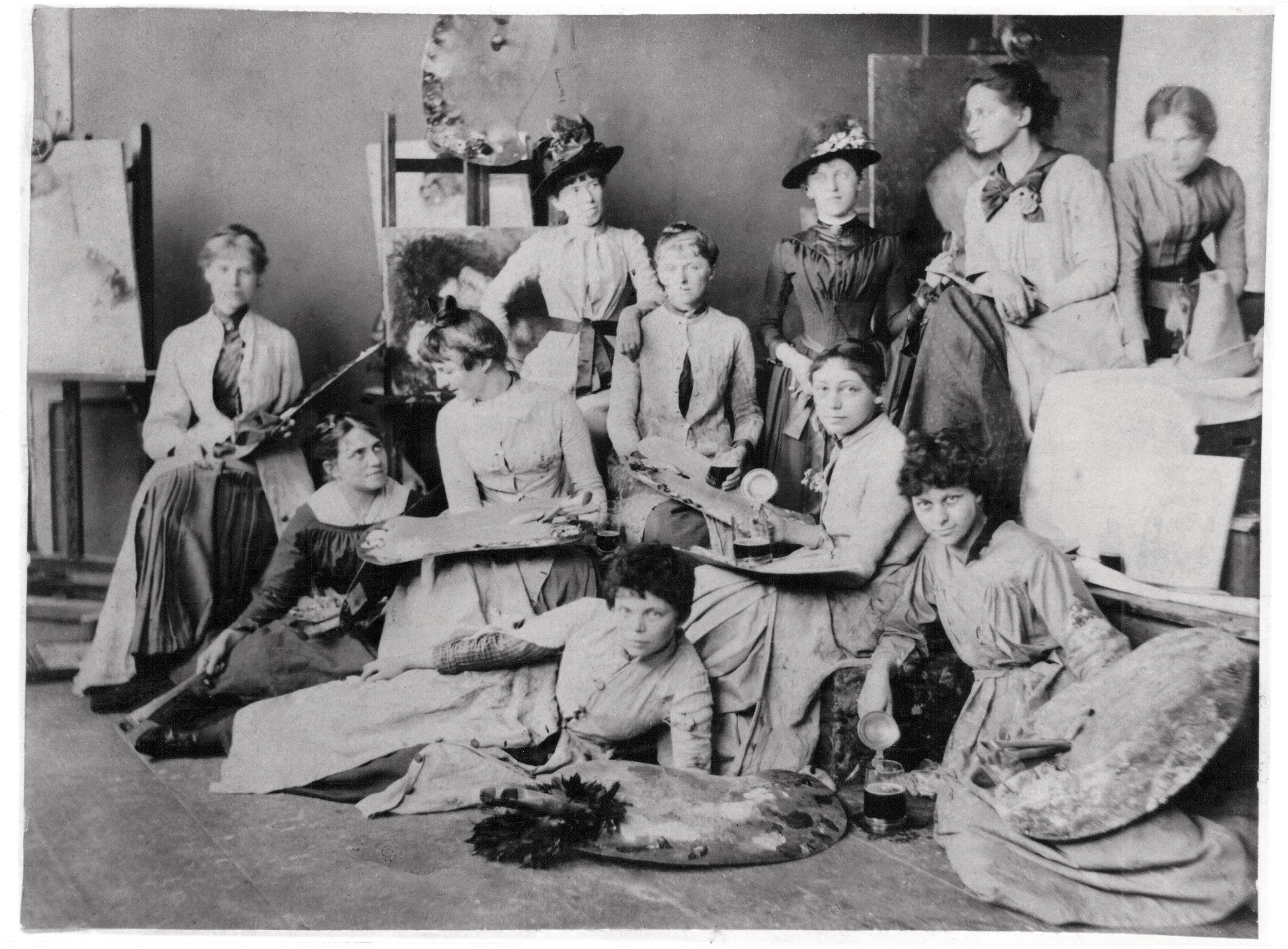 The black-and-white photograph, dating from around 1889, shows a group of eleven young women from Ludwig Herterich’s painting class in Munich, with Käthe Schmidt seated second from the right. The women are arranged in relaxed poses throughout the studio among easels and canvases. They wear high-necked contemporary dresses and some hold artists’ tools such as palettes or, as a humorous detail, a beer stein. The studio appears lively and unconventional, with some of the artists seated on the floor while others stand in the background or perch on stools.