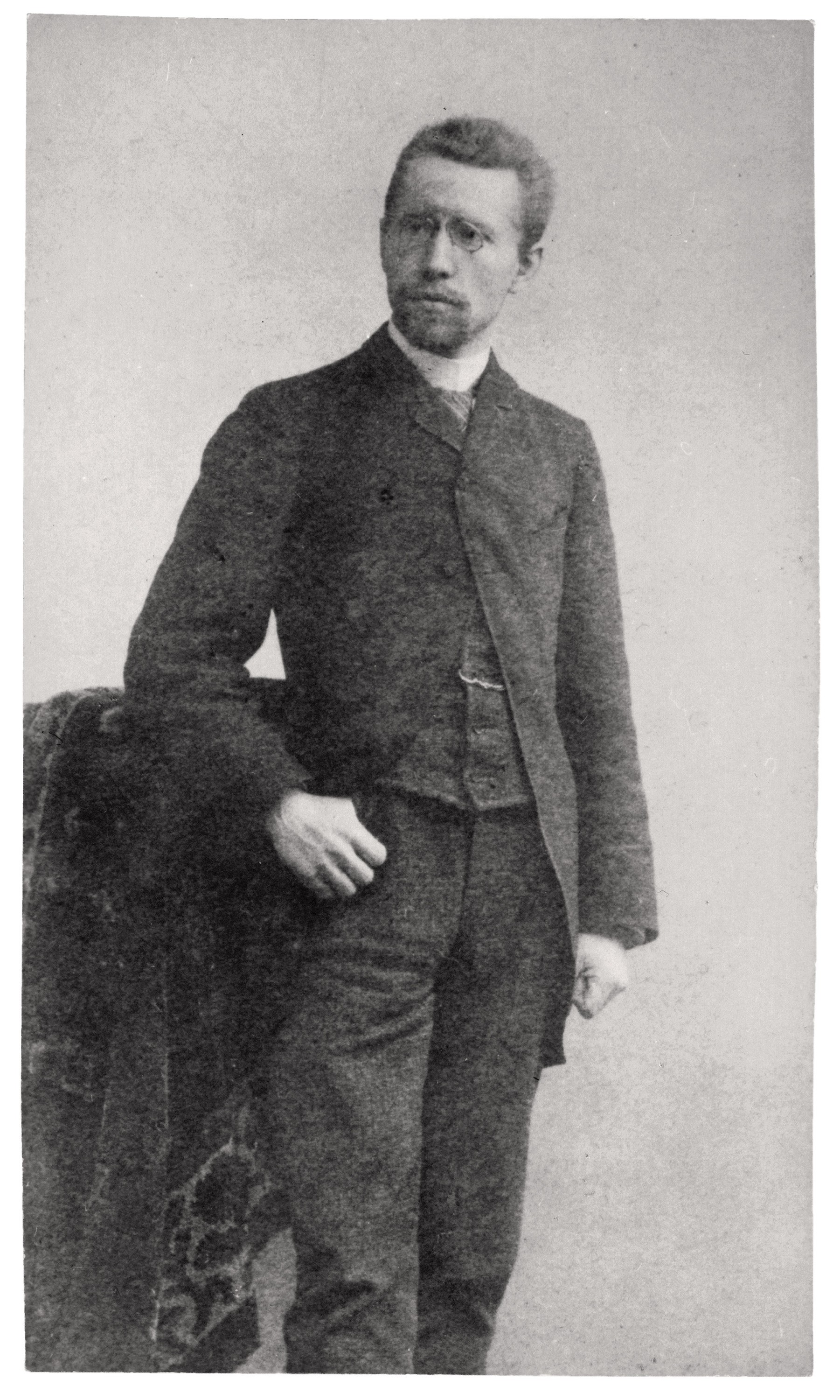 The image shows a historic black-and-white portrait of Karl Kollwitz, dating from around 1885, depicting him as a slender man standing in a formal pose. He wears a dark frock coat over a waistcoat, has a short full beard and spectacles, and rests his right hand casually on his hip. His expression is serious, and his gaze is directed slightly past the viewer into the distance as he stands before a plain, light-coloured background.